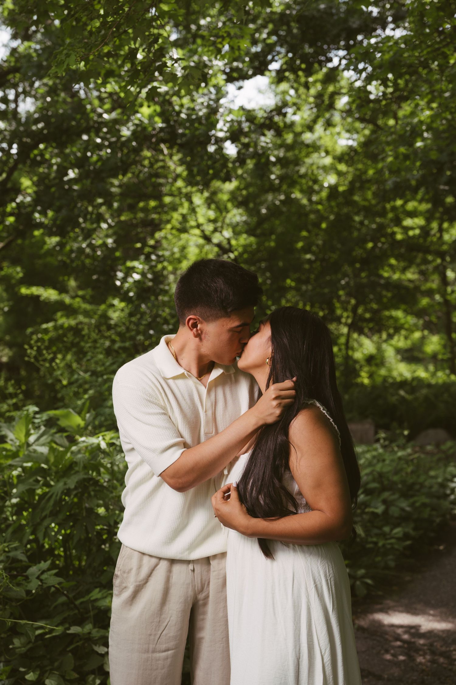 Couple sharing a tender kiss surrounded by lush green trees in a sunlit park setting.