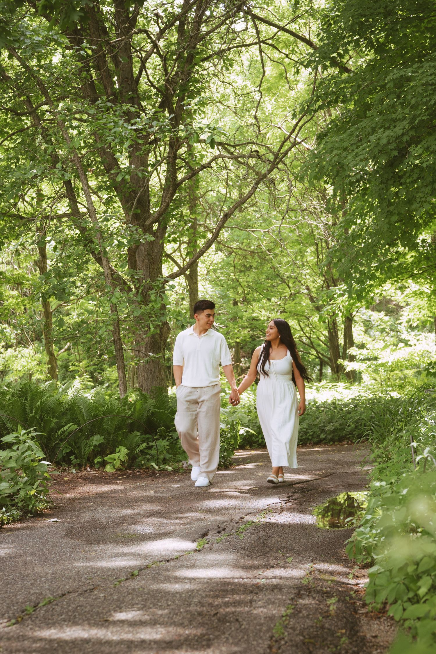 Couple holding hands walking along a lush green tree-lined path wearing matching white outfits.
