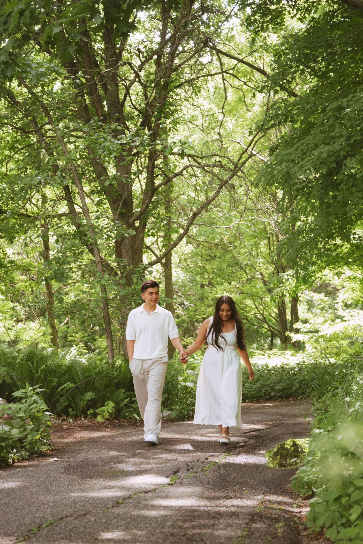 Couple holding hands walking on a tree-lined path in a lush green park, both dressed in white outfits.