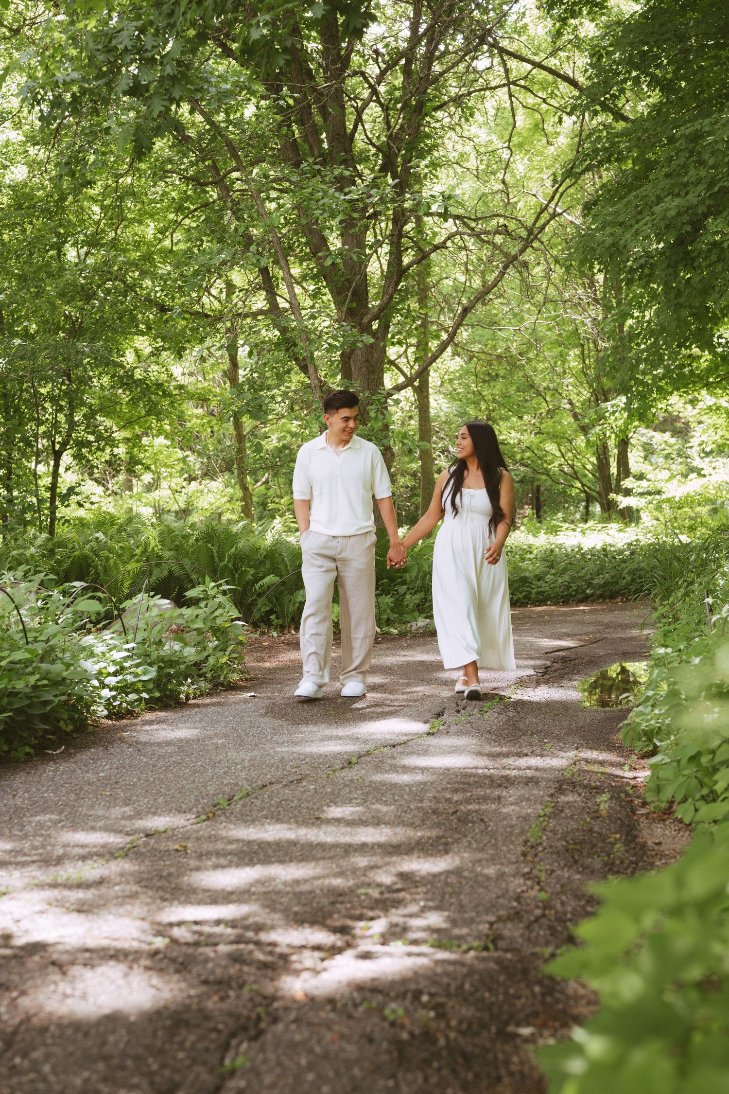 A couple walks hand in hand along a sunlit garden path surrounded by vibrant green trees and foliage.