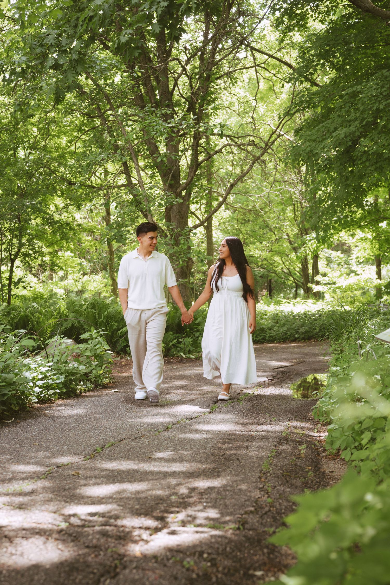 Couple holding hands and walking along a sunlit tree-lined garden path, both dressed in white and beige attire.
