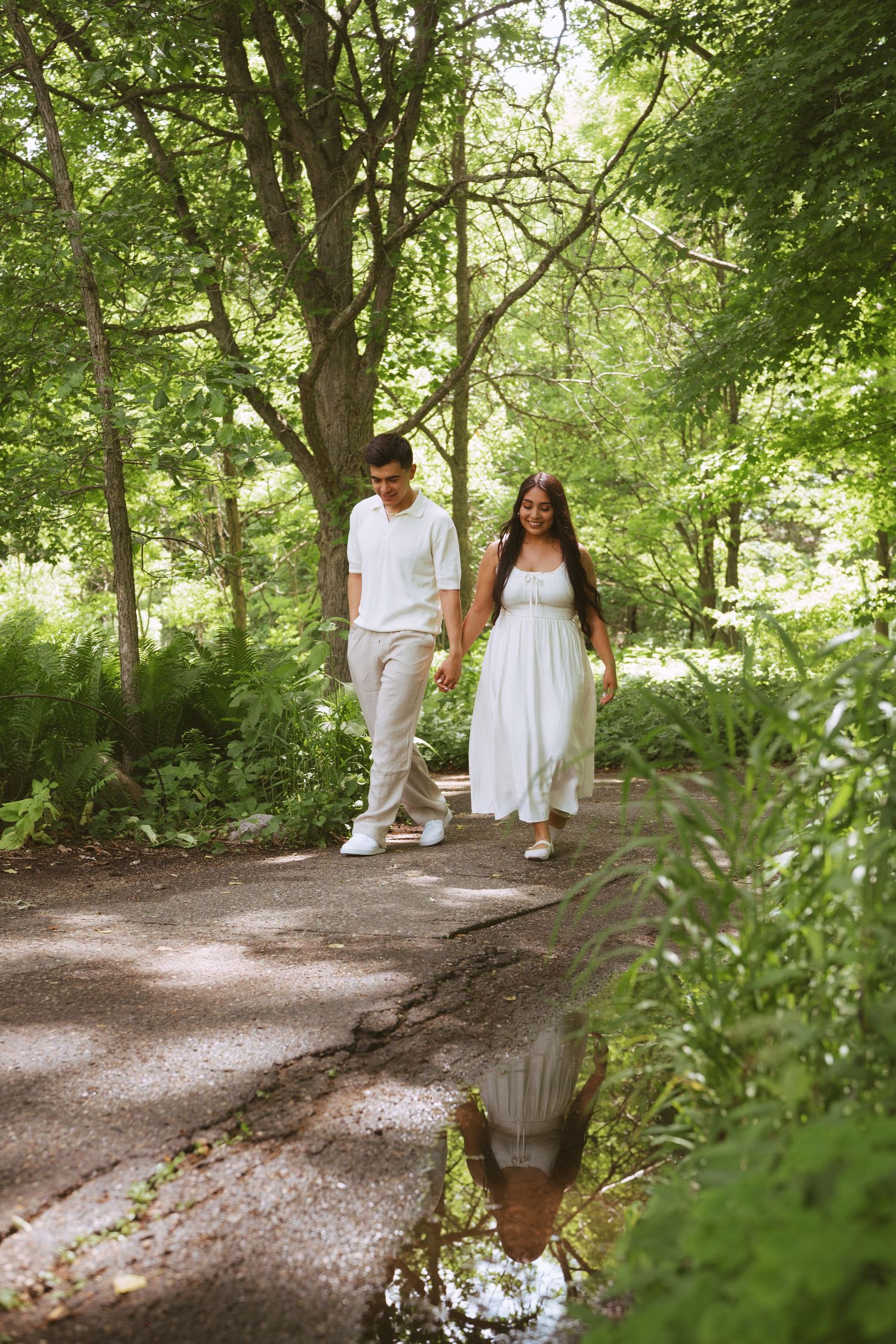 Couple in white outfits holding hands while walking on a lush green park path with puddle reflection.