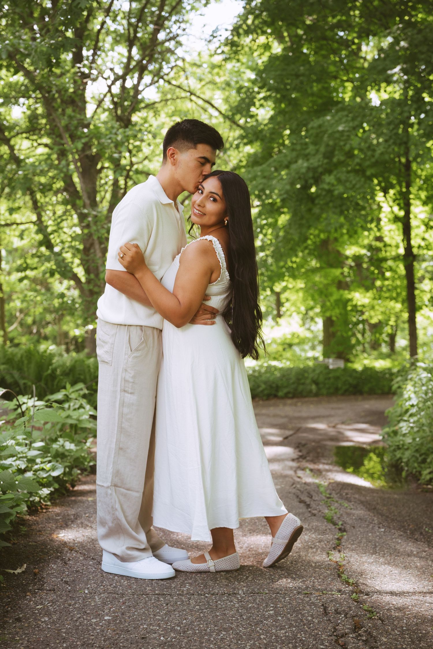 Couple embracing on a sunlit park path, woman in white dress smiling, man in beige outfit, surrounded by lush green trees.