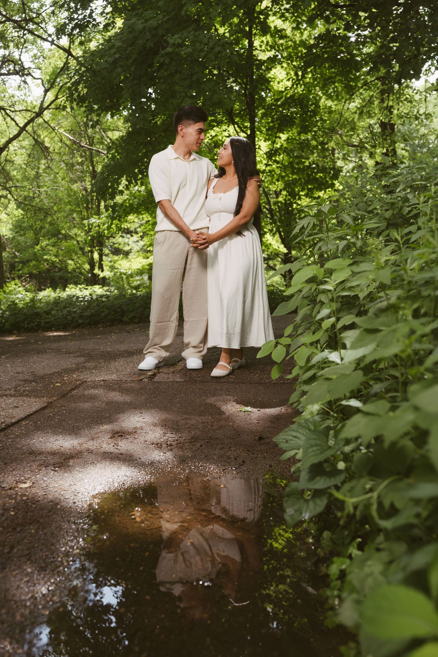 Couple holding hands and gazing at each other on a garden path with a puddle reflection.