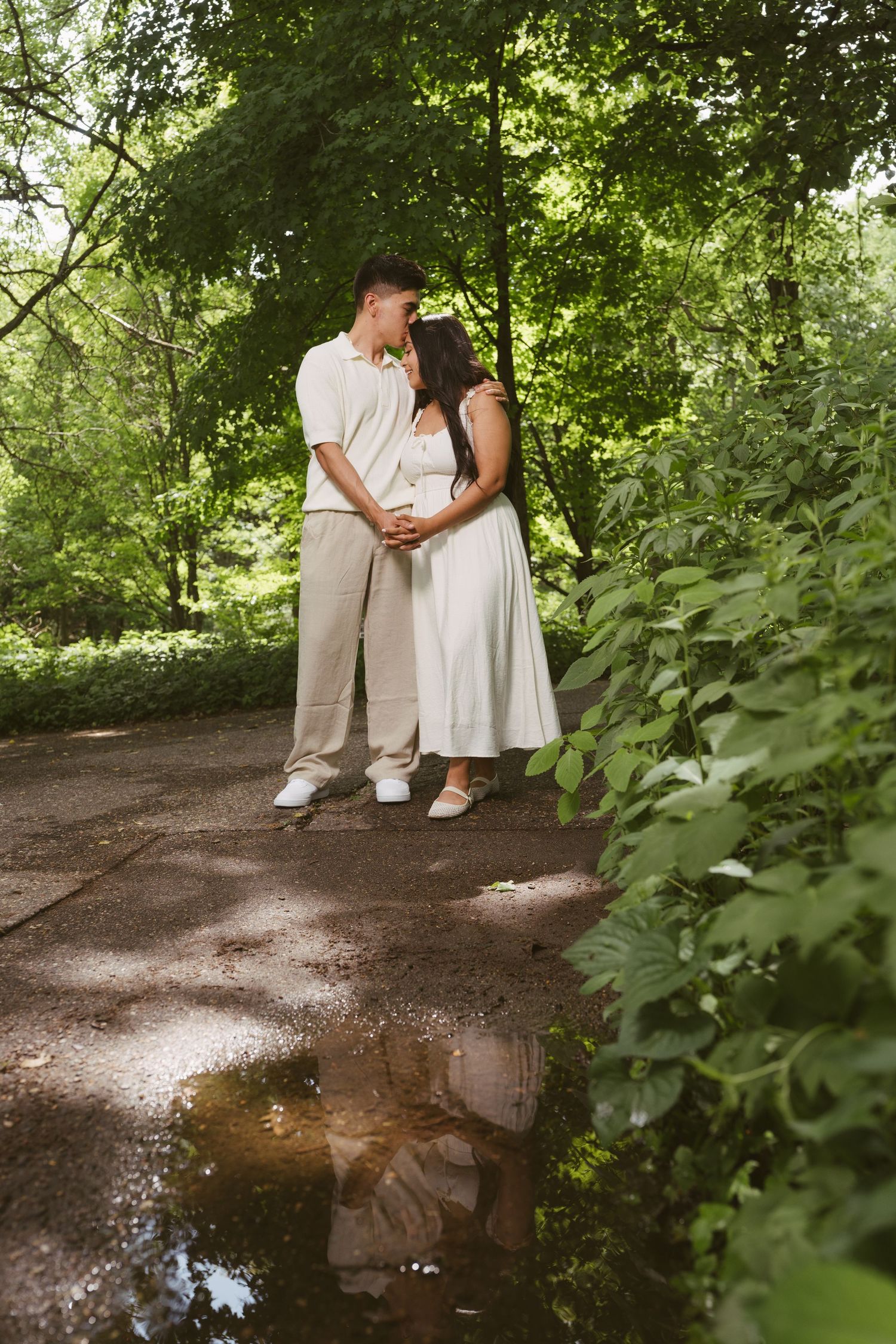 Romantic couple in white clothing standing close together on a tree-lined park path near a puddle.