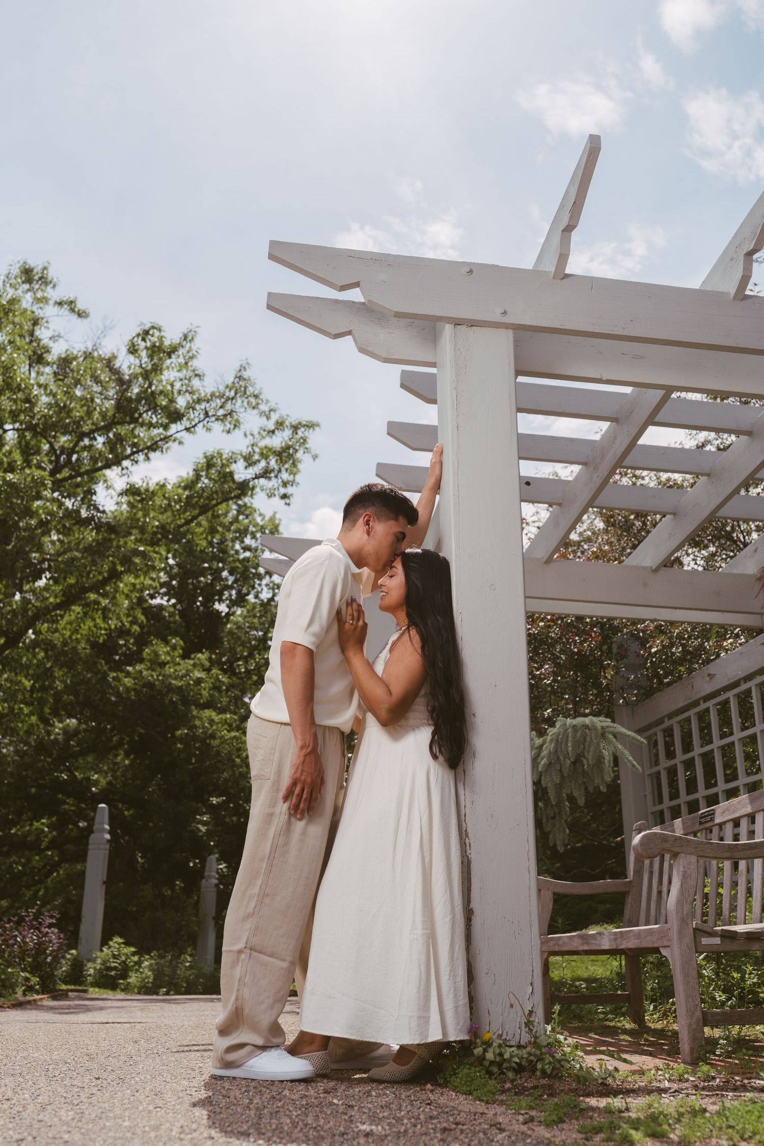 Couple sharing a kiss under a white pergola in a sunny garden setting with blue sky.