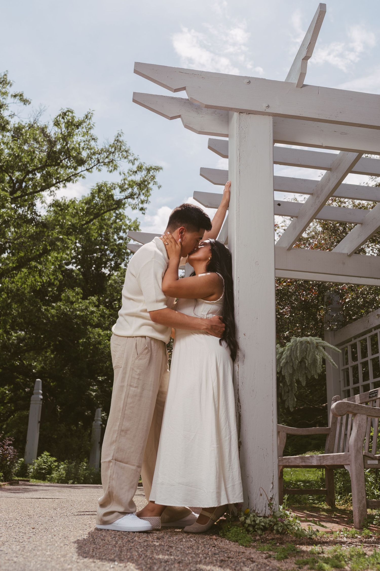 Couple sharing a romantic kiss under a white wooden pergola in a sunny garden setting.