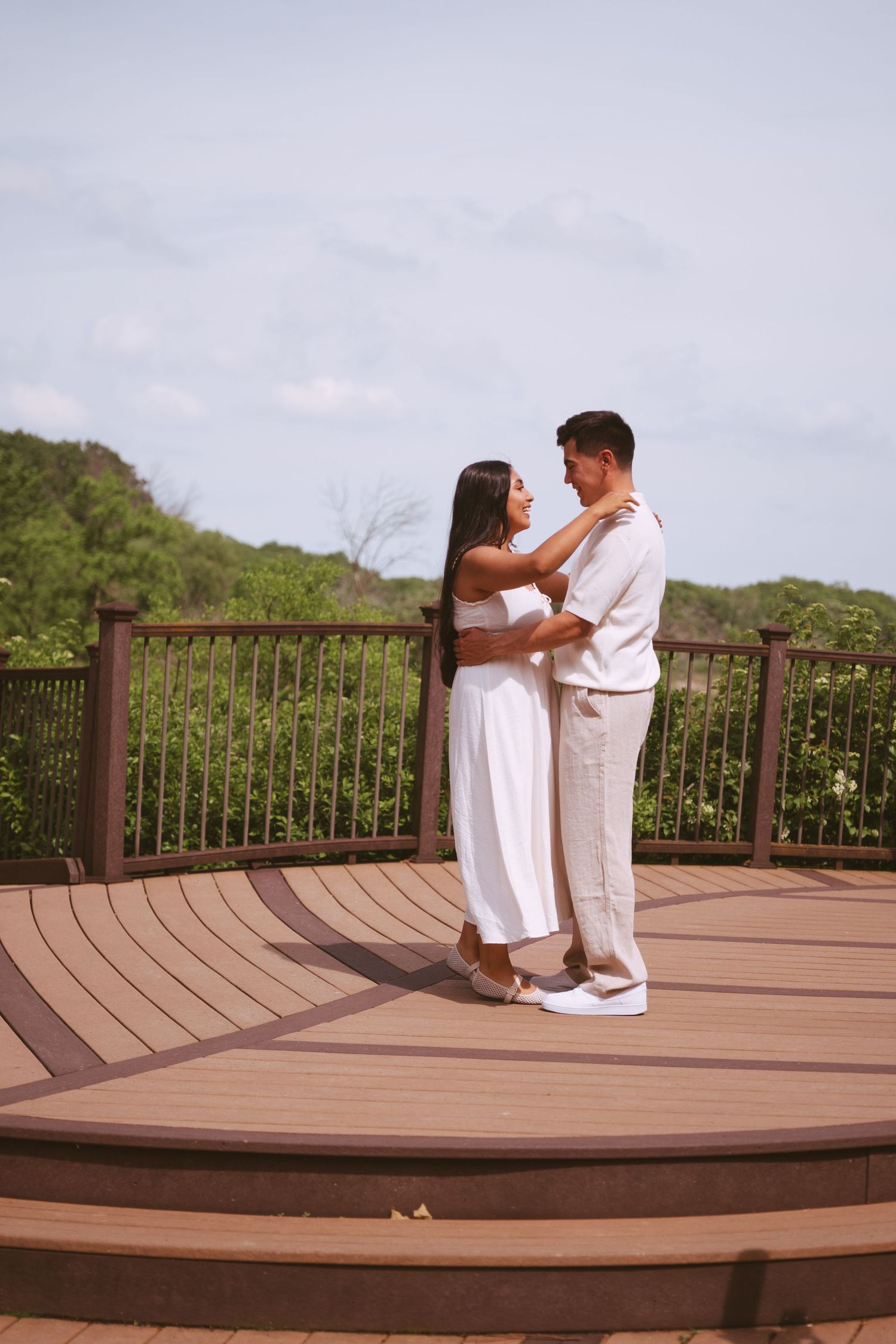 Couple in white outfits embracing on a wooden deck overlooking lush green trees under a blue sky.