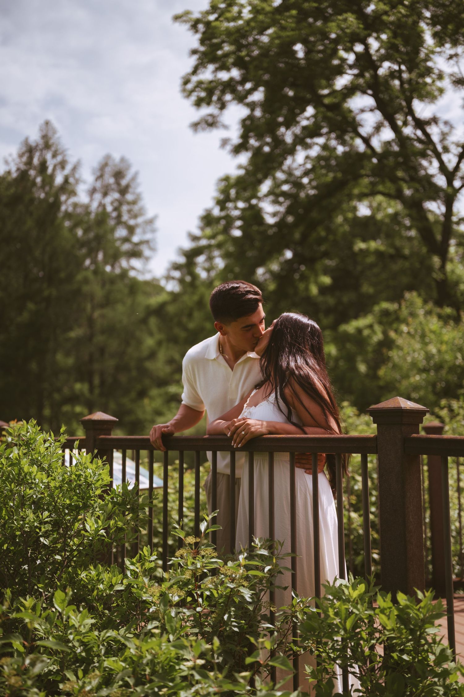 Couple sharing a romantic kiss on a wooden deck surrounded by lush green trees and shrubs outdoors.