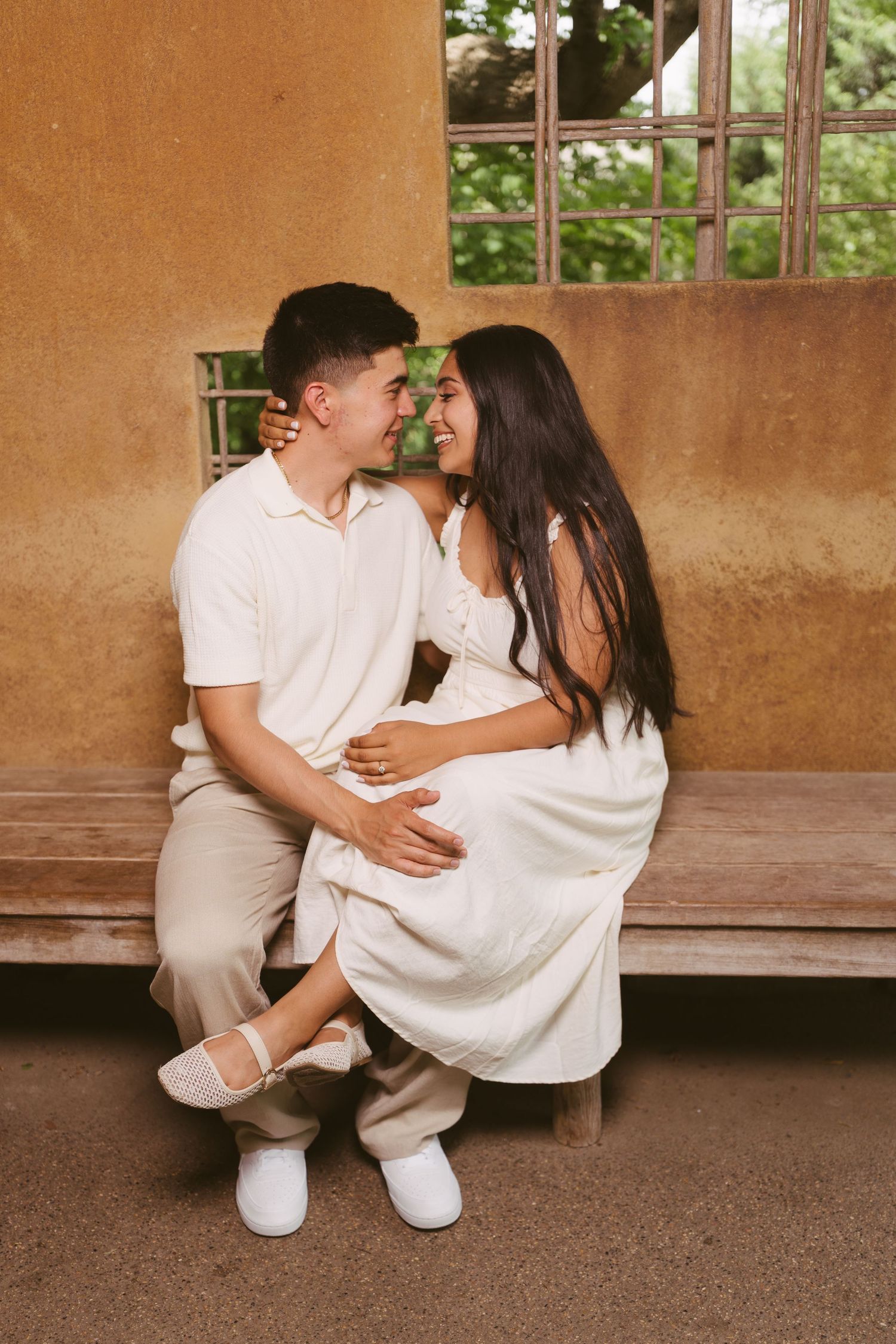 A couple in white outfits sits on a bench, smiling at each other in a warm, romantic outdoor setting.