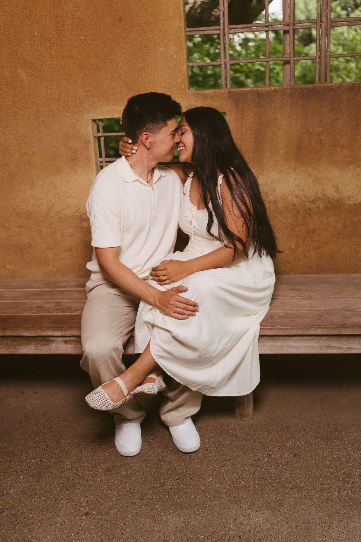 A couple in matching white outfits share a romantic moment on a wooden bench against a warm adobe wall.