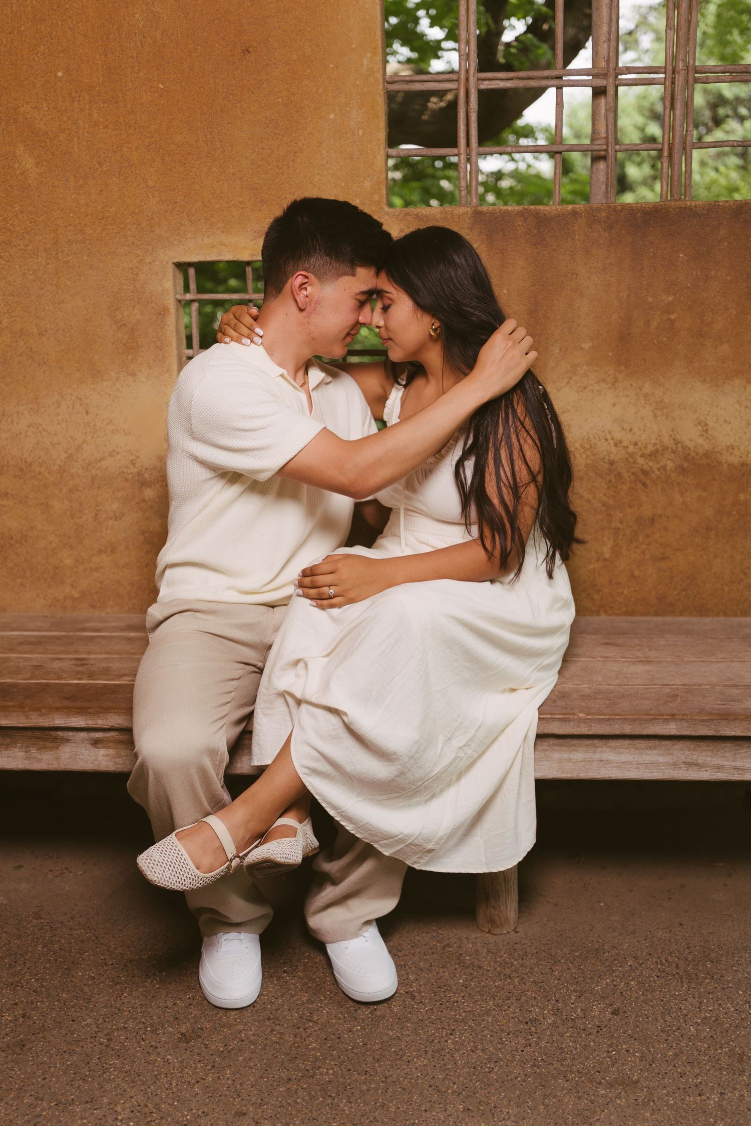 Couple in matching cream outfits sitting close together on a wooden bench, sharing an intimate moment outdoors.