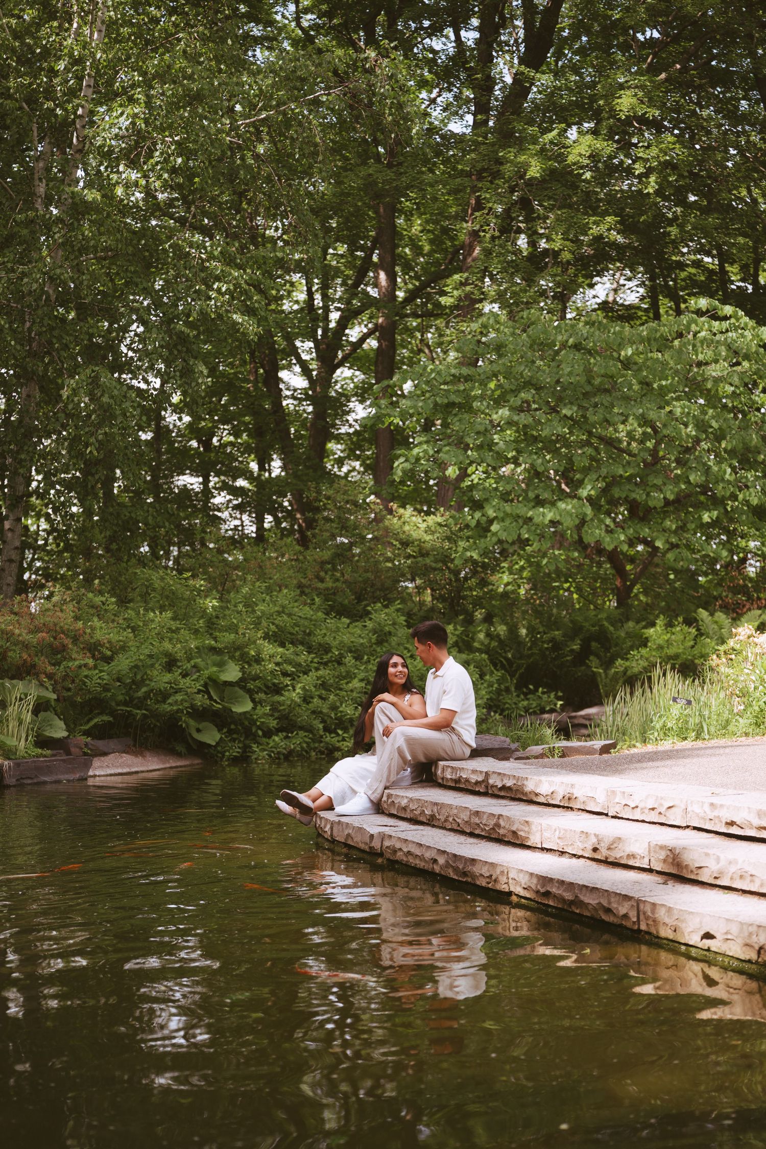 Couple sitting closely on stone steps beside a serene pond surrounded by lush green trees.