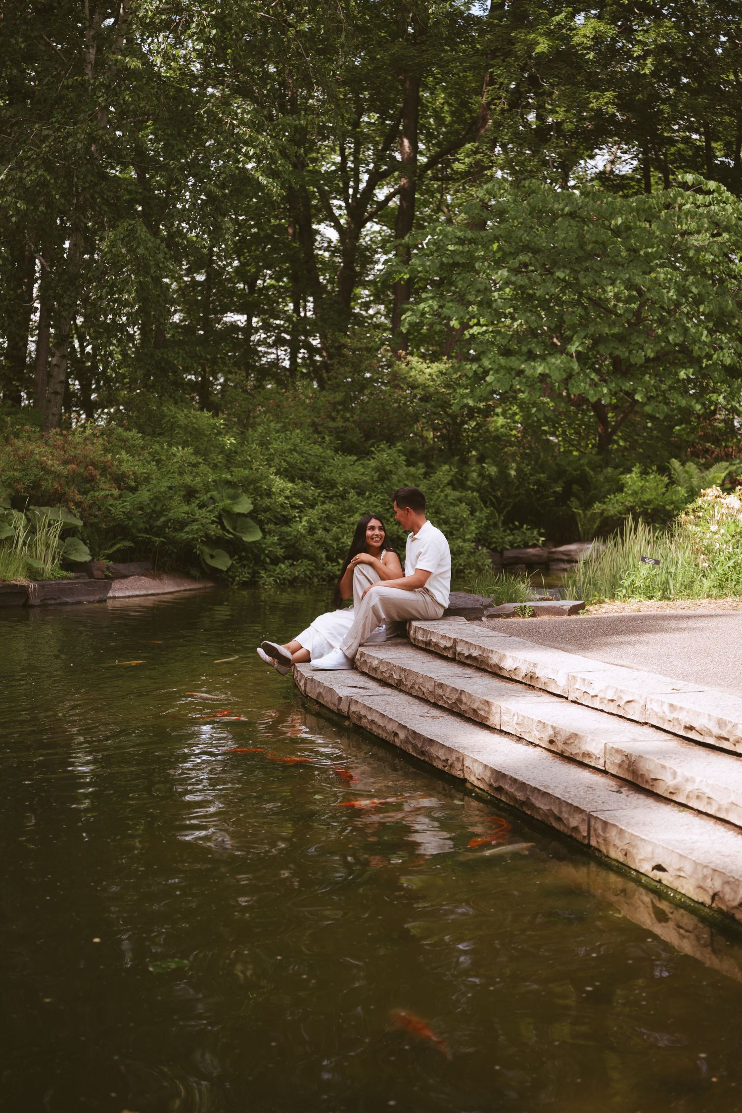 Romantic couple sitting on stone steps beside a koi pond in a serene green garden.