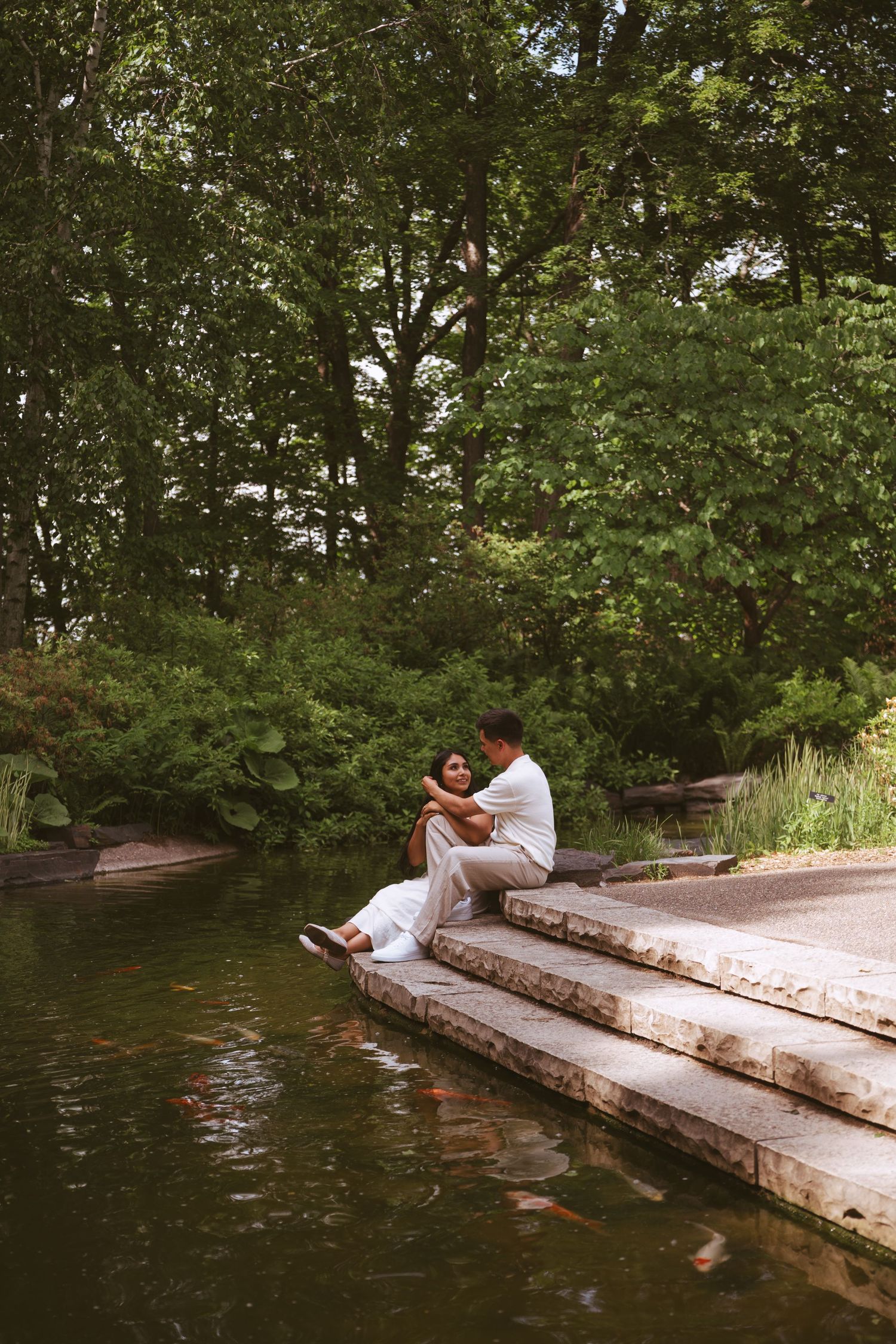 Couple sitting close together on stone steps beside a koi pond in a serene green park.