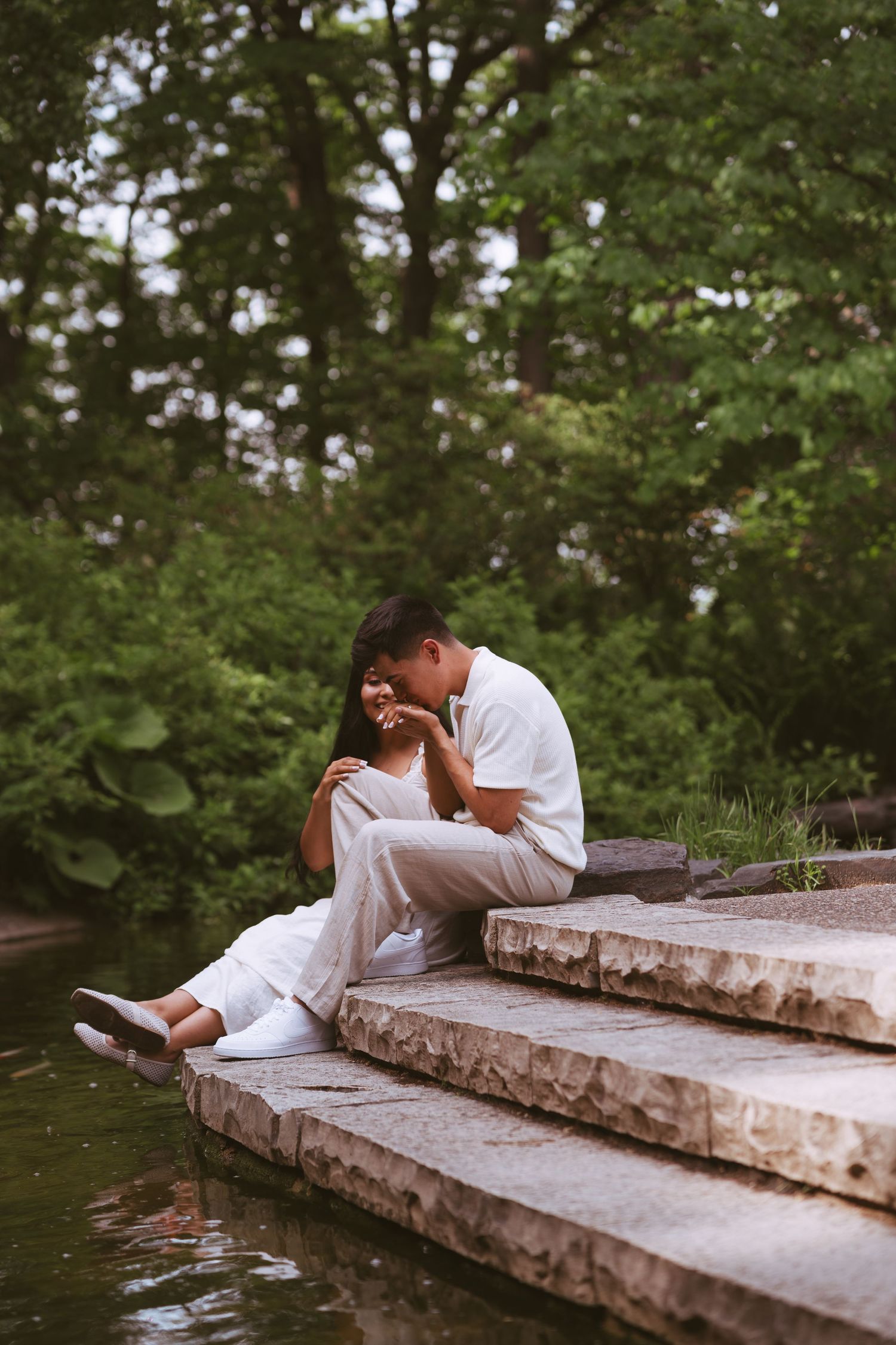 Couple sitting on stone steps by a pond in a green park, man kissing woman's hand.