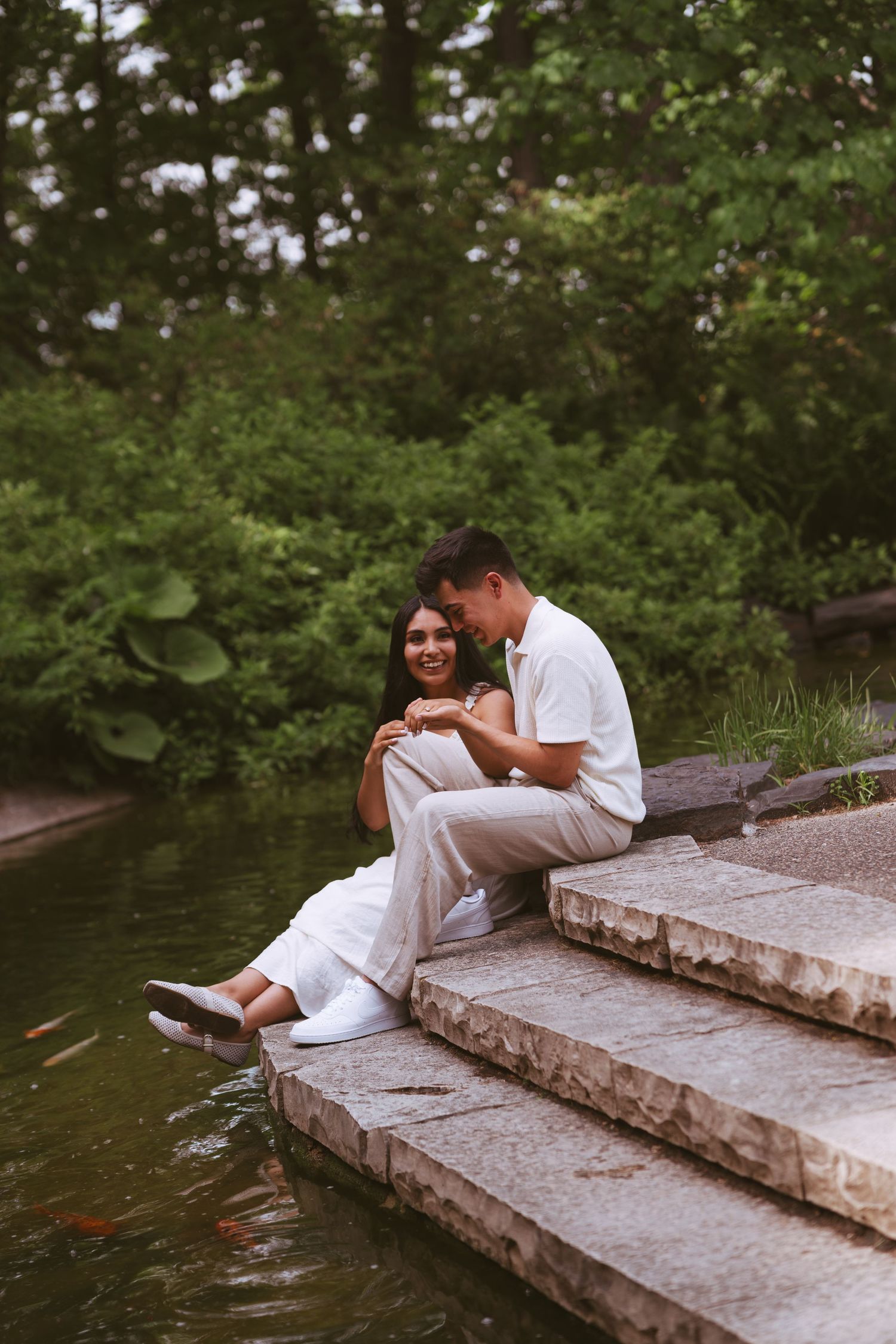 Happy couple sitting on stone steps by a koi pond, surrounded by lush greenery.