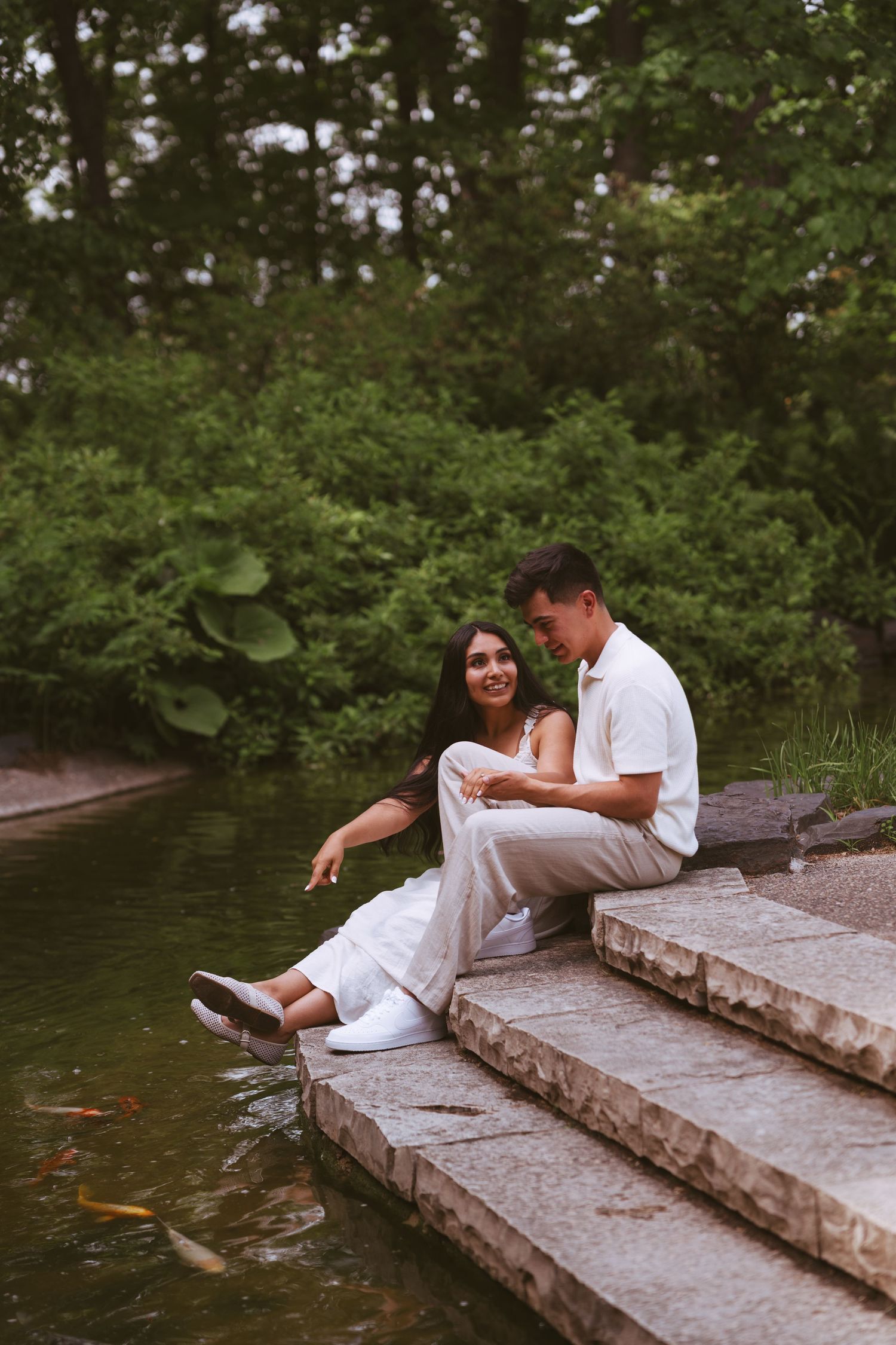 Smiling couple relaxing on stone steps by a koi pond, surrounded by lush greenery.