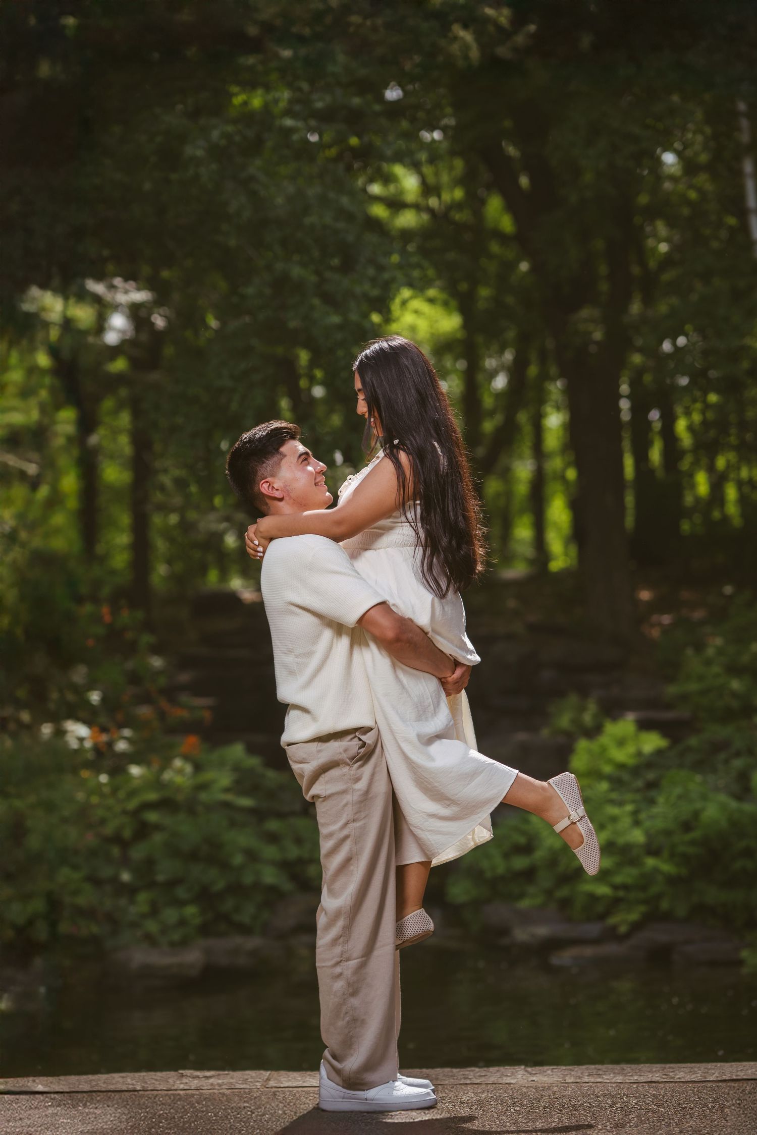 Man lifting woman in white dress in a lush green park, both smiling and gazing at each other lovingly.