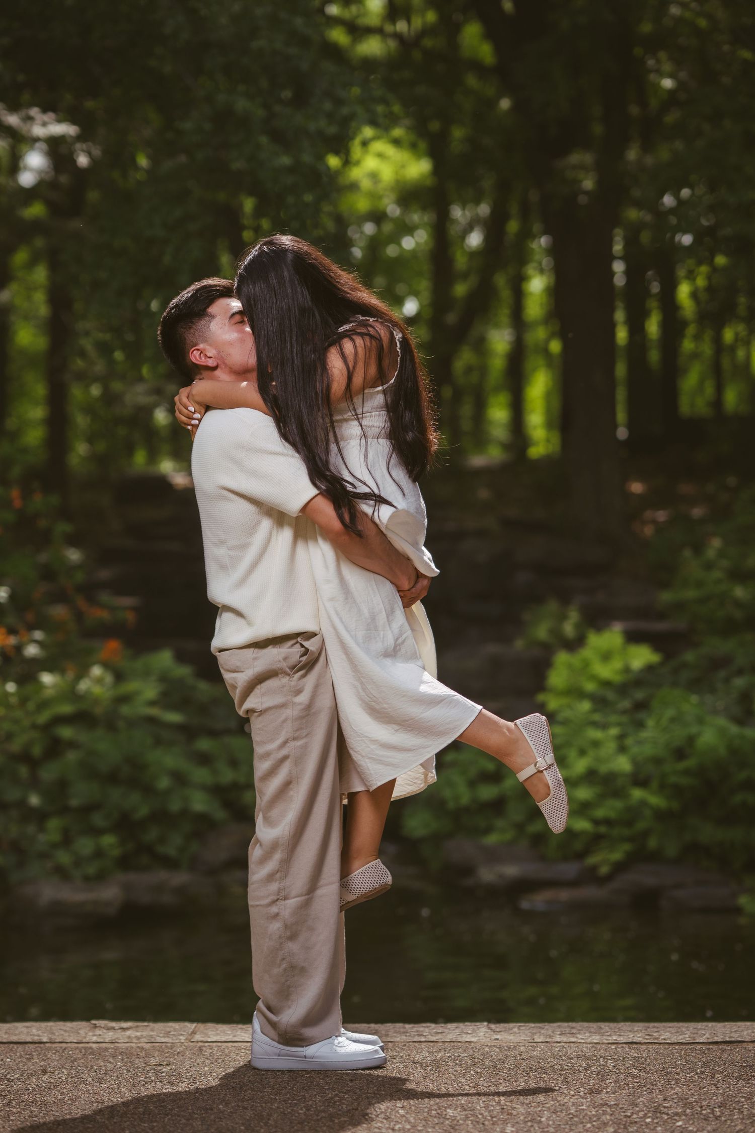 Man lifting woman in a joyful embrace in a sunlit park, both wearing white and beige outfits.