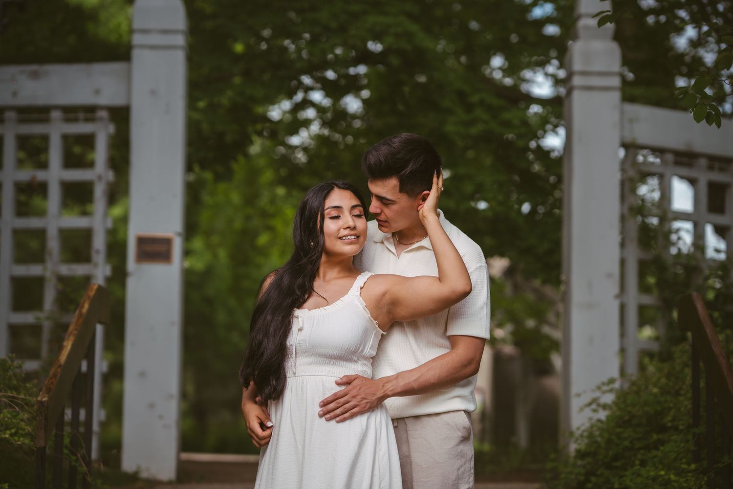 Couple embracing near a white pergola in a lush garden, woman in white dress smiling softly.