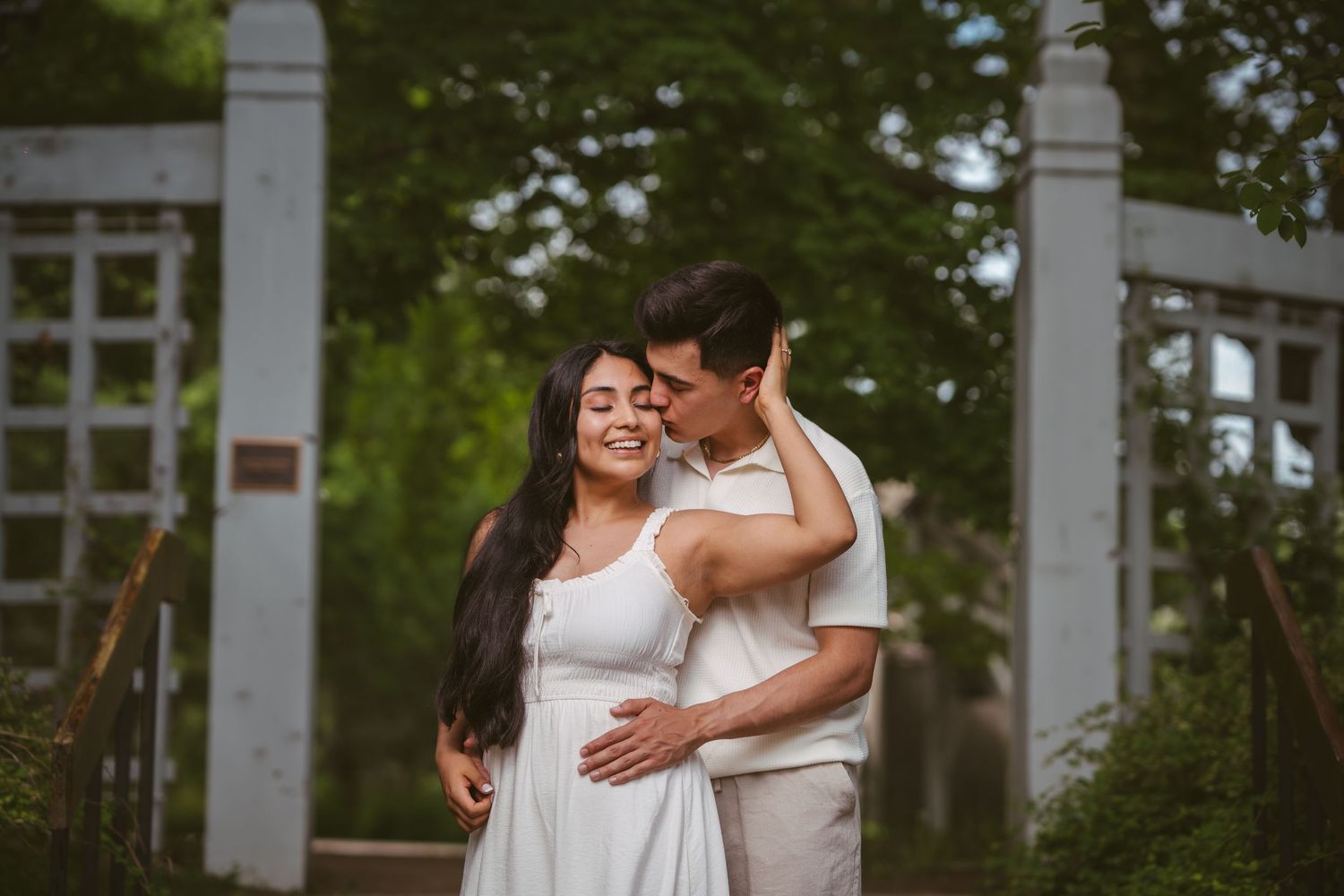 Couple embracing near white garden arbor, woman in white dress smiling joyfully.