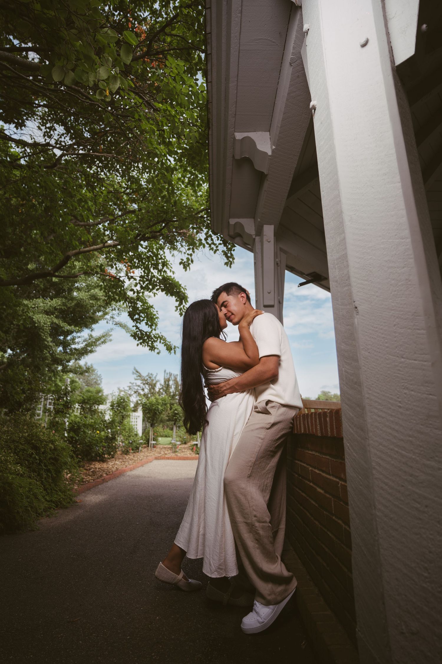 Couple sharing a romantic kiss leaning against a brick building corner surrounded by green trees.