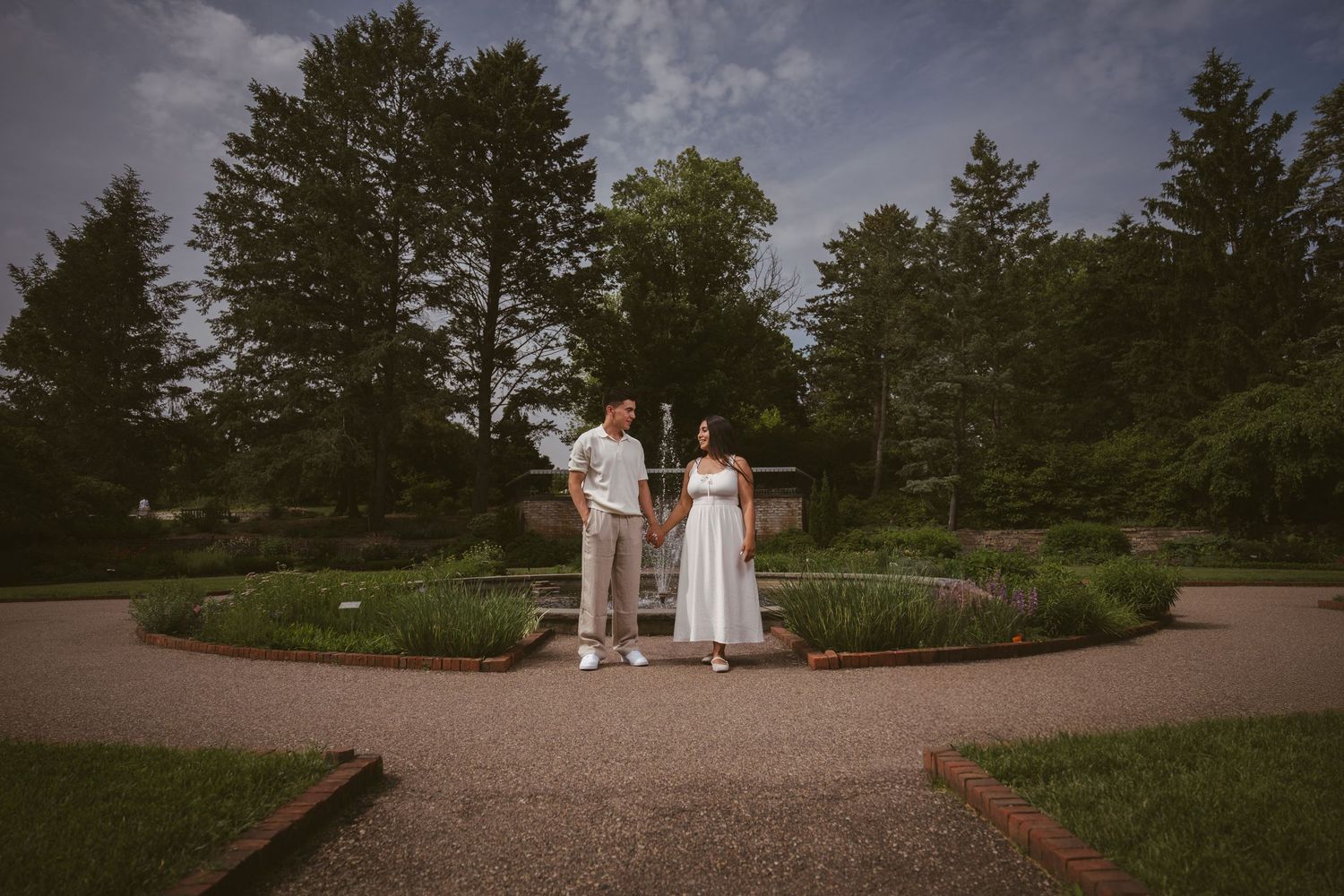 Engaged couple holding hands standing in a formal garden with a white pergola and lush trees.