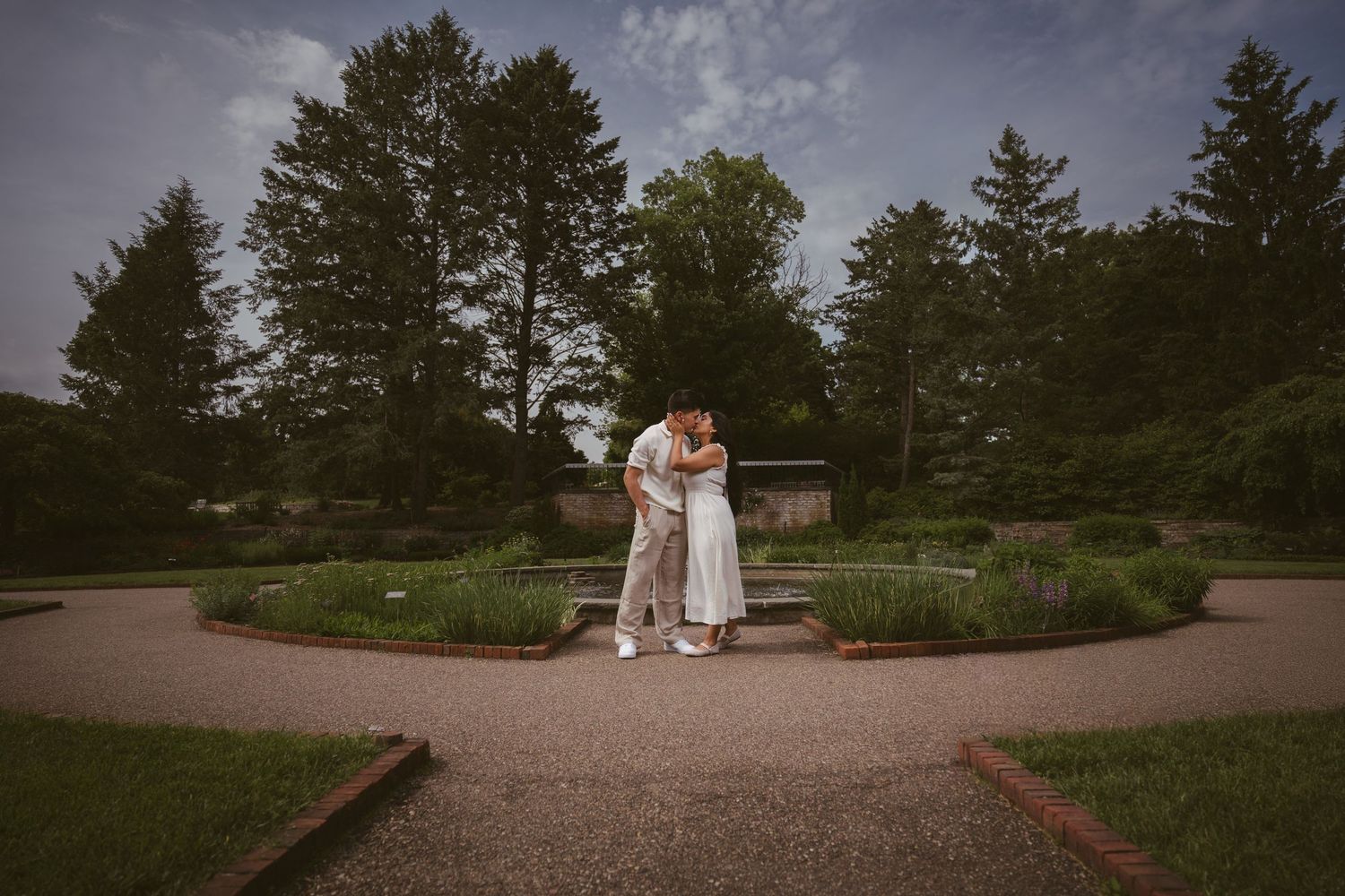 A couple shares a romantic embrace in the center of a circular garden path surrounded by lush greenery.