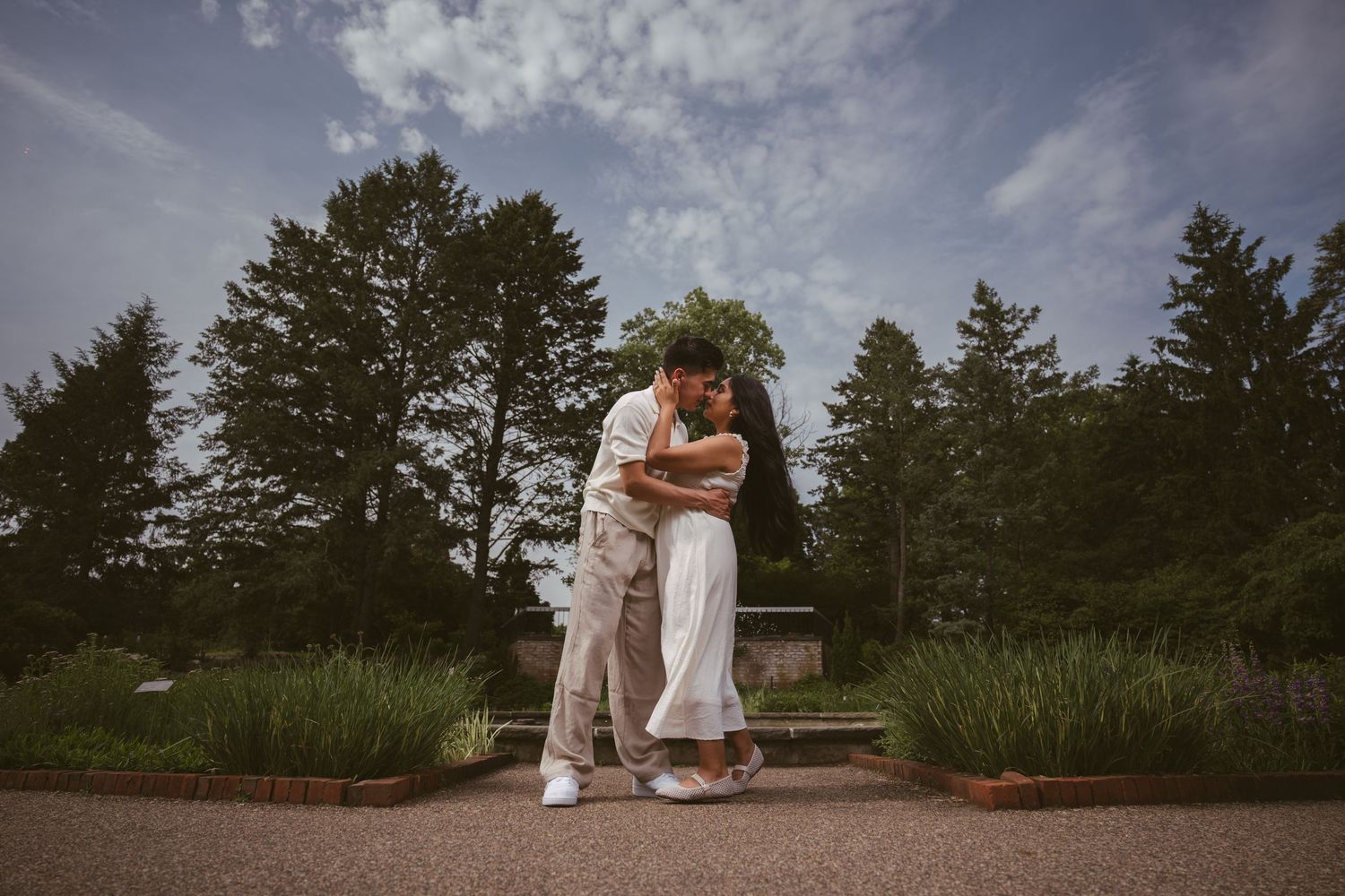 Couple sharing a tender embrace on a garden path with tall trees and blue sky in the background.