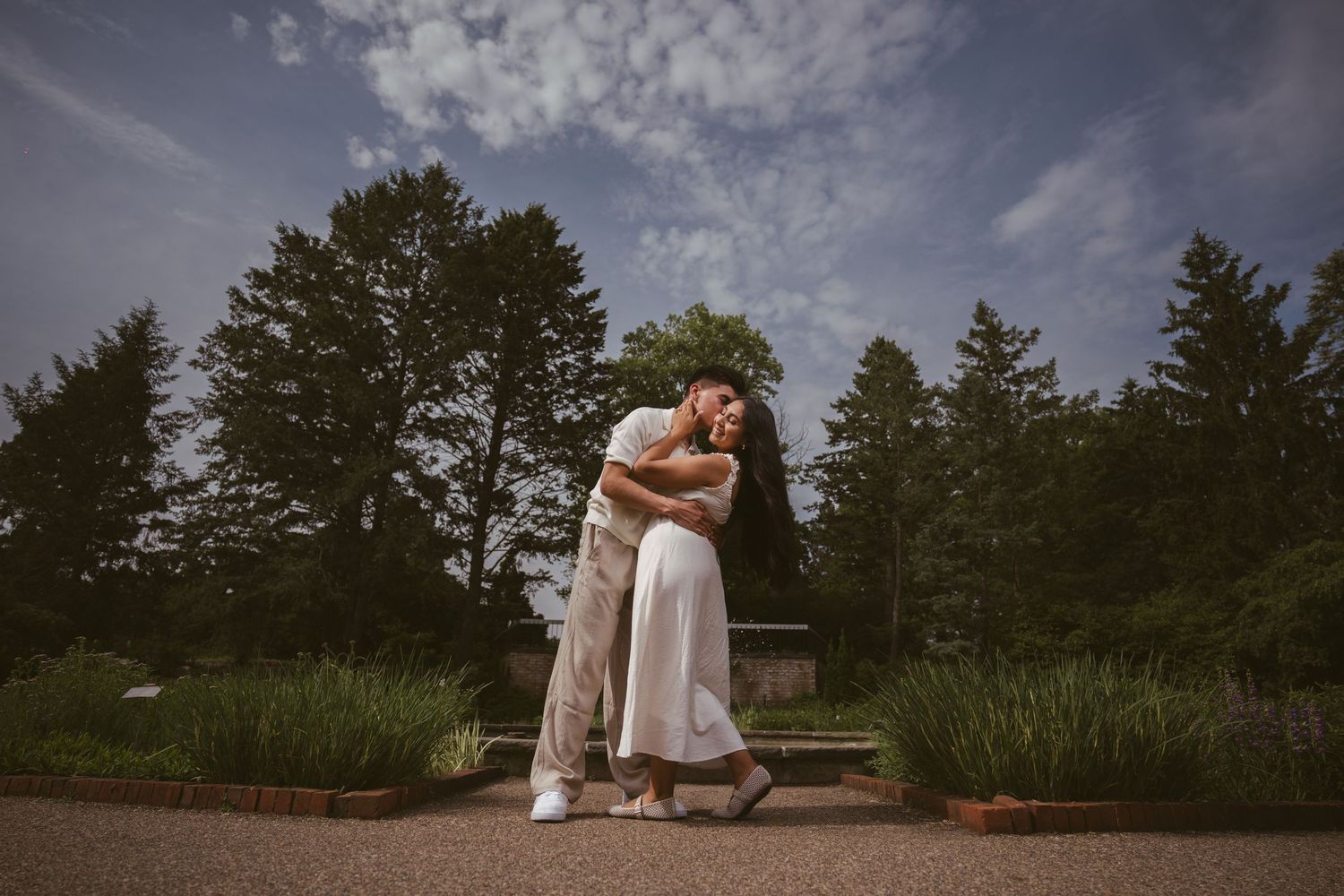 Couple sharing a romantic embrace and dip in an outdoor garden under a dramatic blue sky.