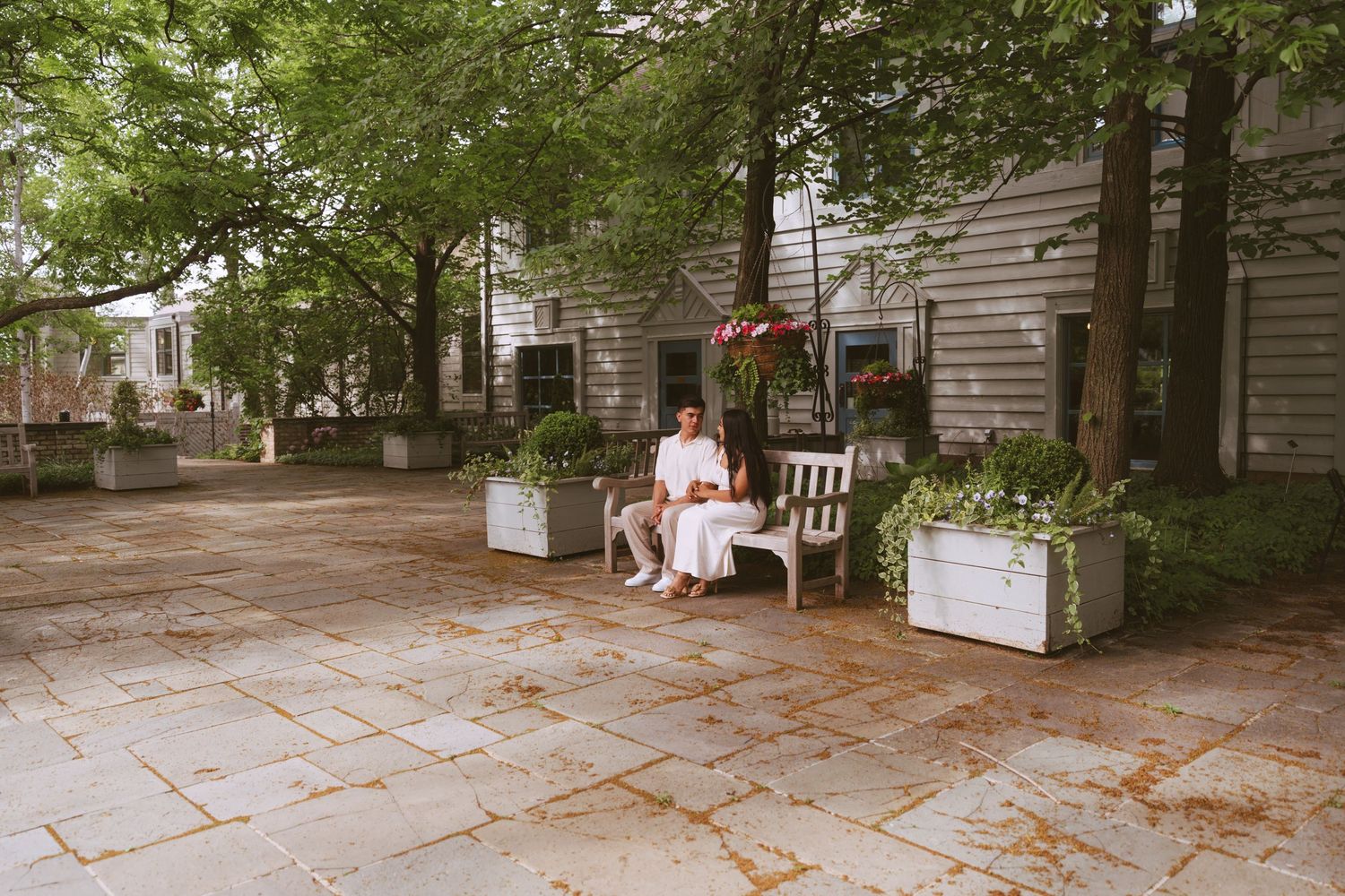 Woman in white dress sitting alone on a bench in a charming tree-lined courtyard setting.
