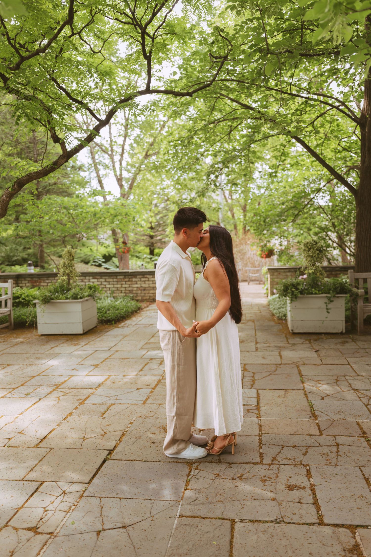 Couple sharing a kiss under a canopy of trees on a stone pathway in a formal garden.