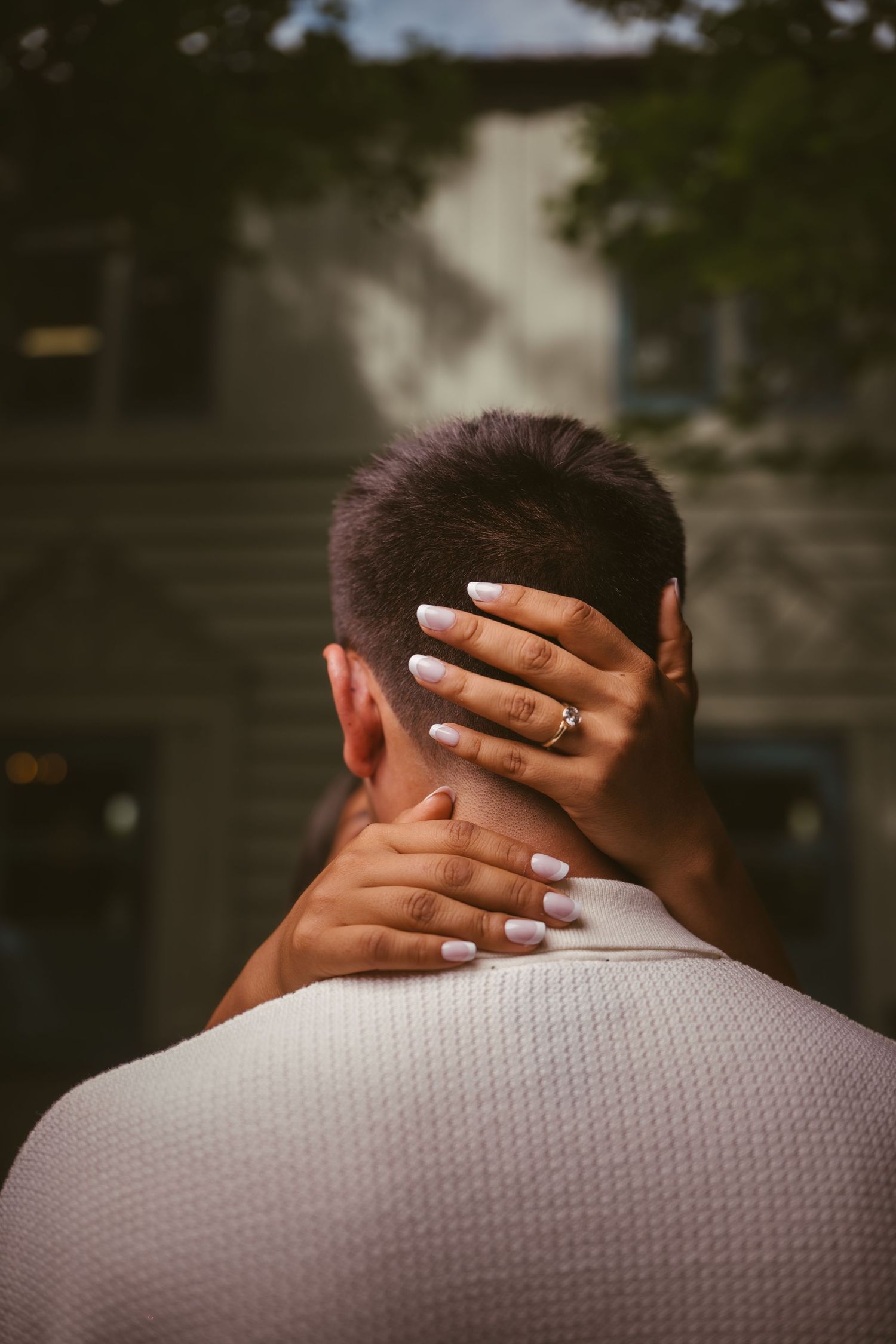 Close-up of woman's hand with engagement ring resting on man's neck from behind.
