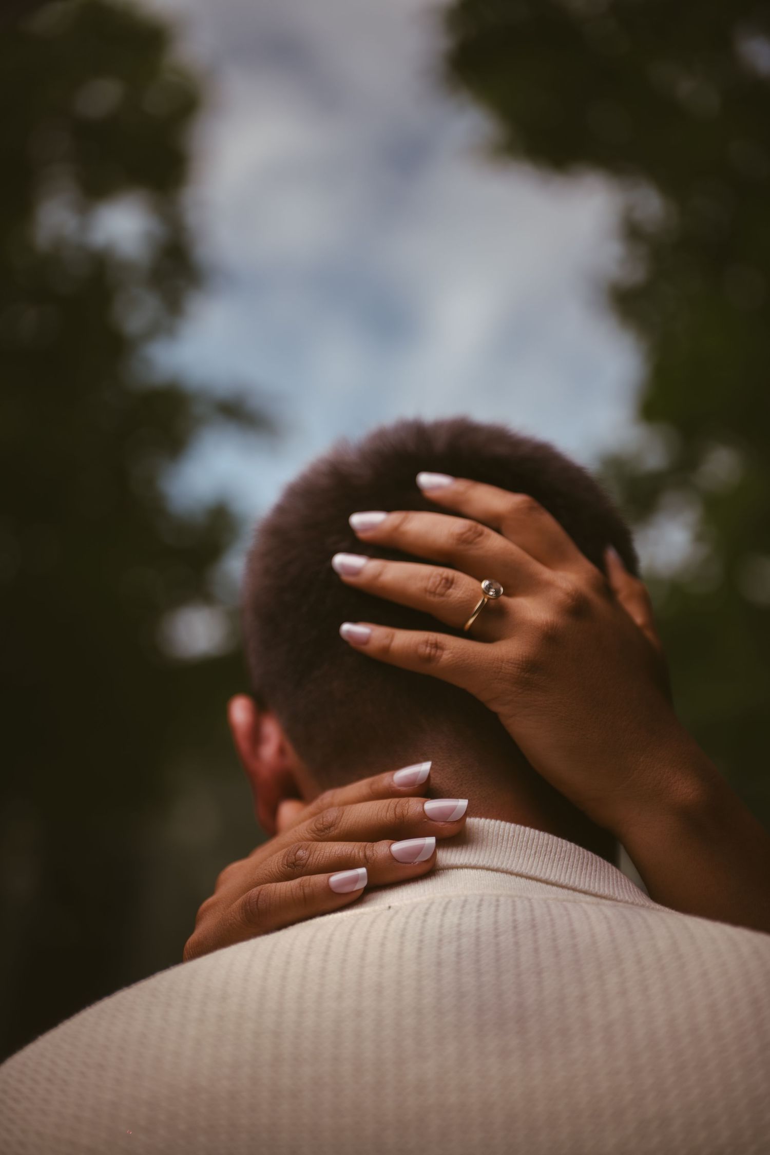 Close-up of a woman's hand with white nails and gold ring resting on a man's head and shoulder outdoors.