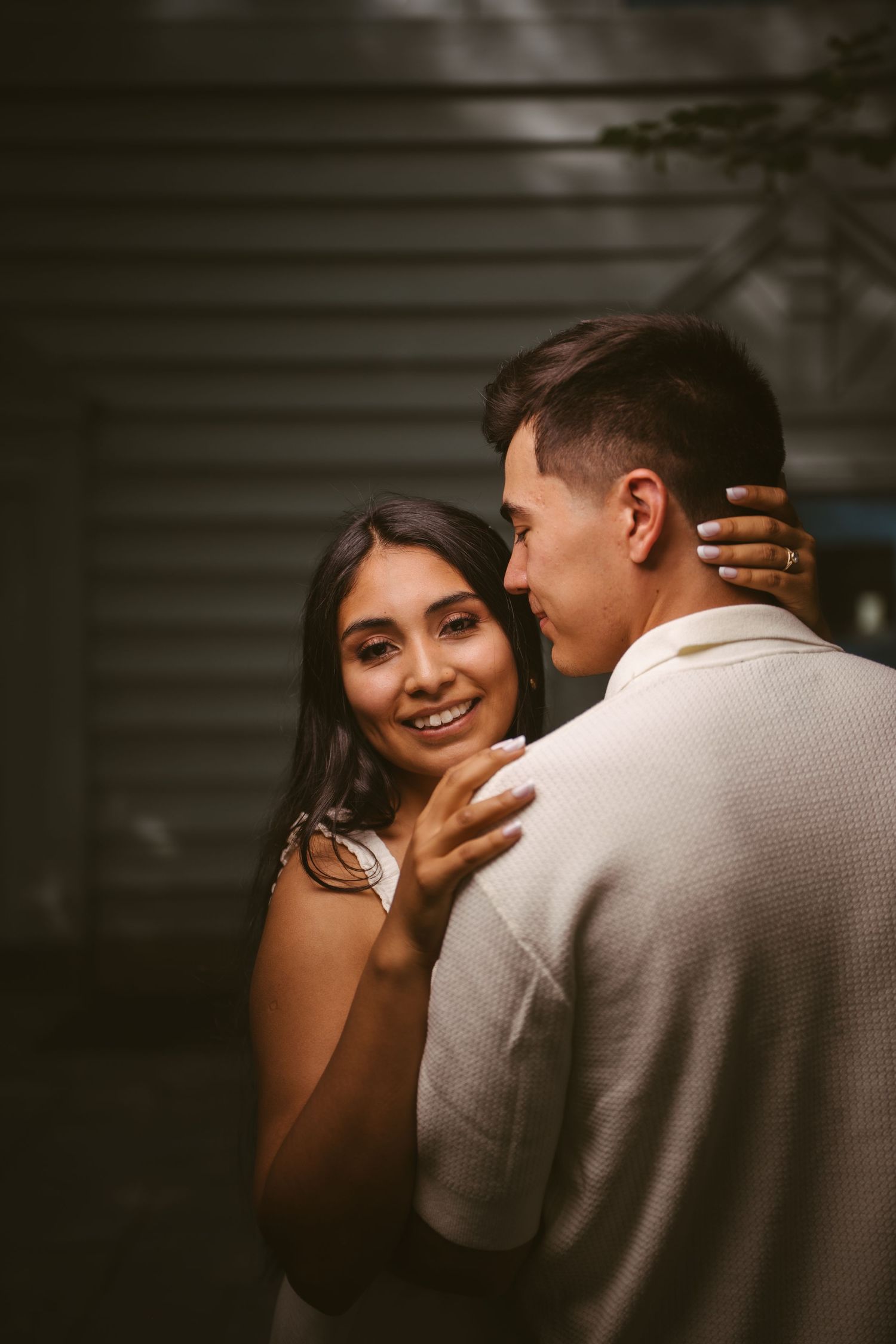 Woman smiling warmly while embracing a man in a beige shirt against a dark background in a romantic portrait.