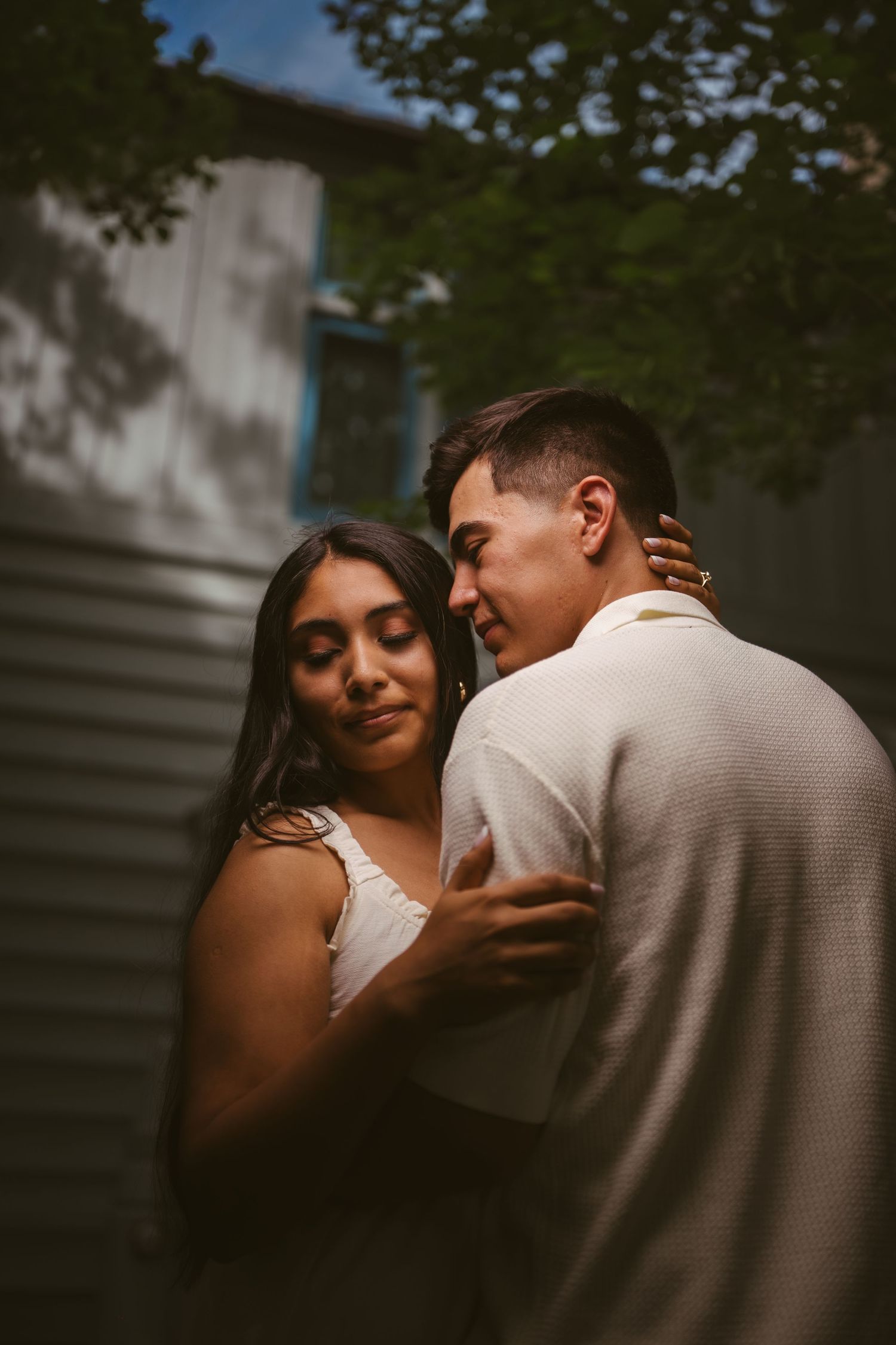 Couple embracing tenderly in soft evening light with trees and a building in the background.
