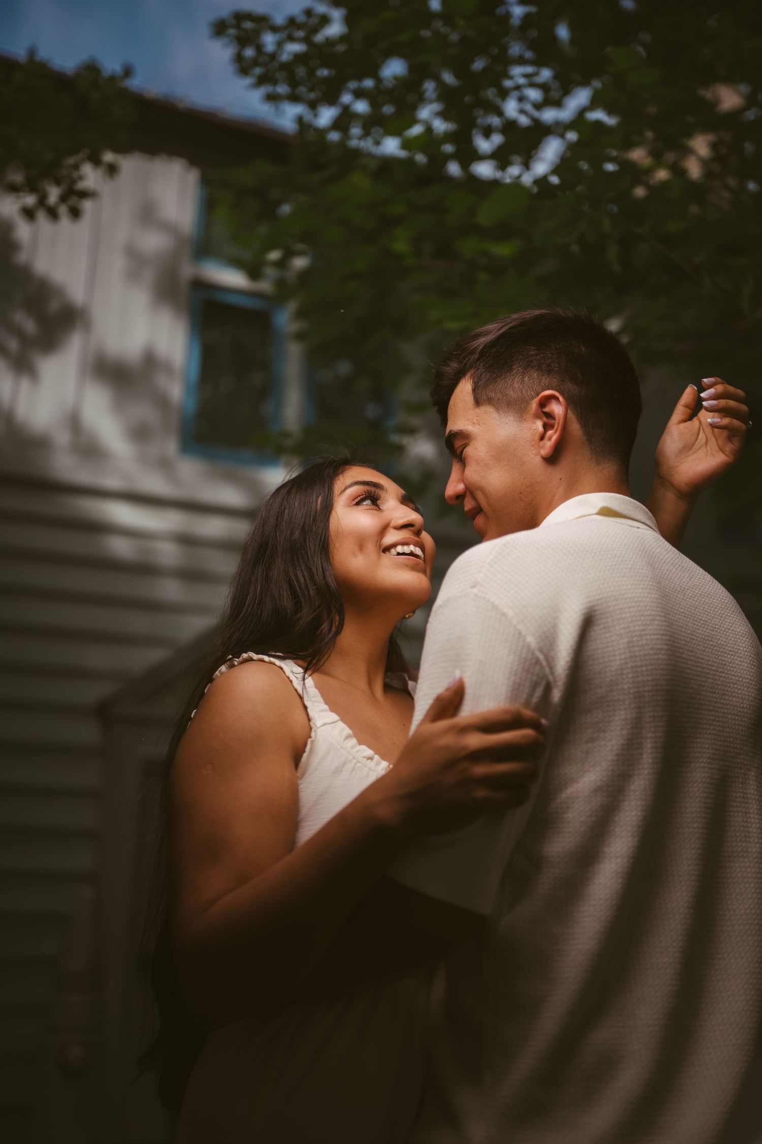 A smiling couple embraces outdoors near a building, sharing a romantic moment surrounded by lush green trees.