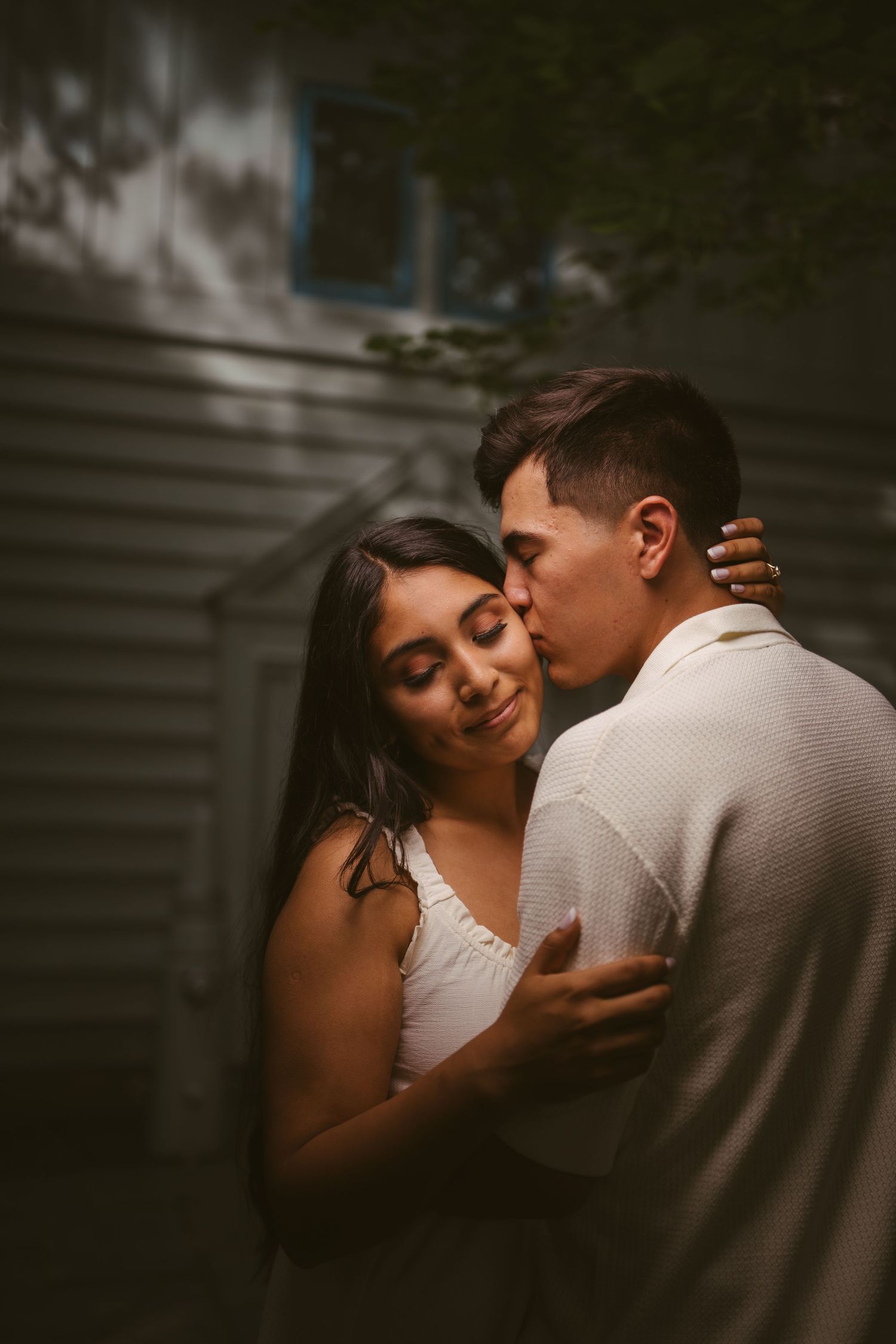 A couple shares a tender kiss in moody outdoor lighting, the woman smiling softly as the man kisses her cheek.