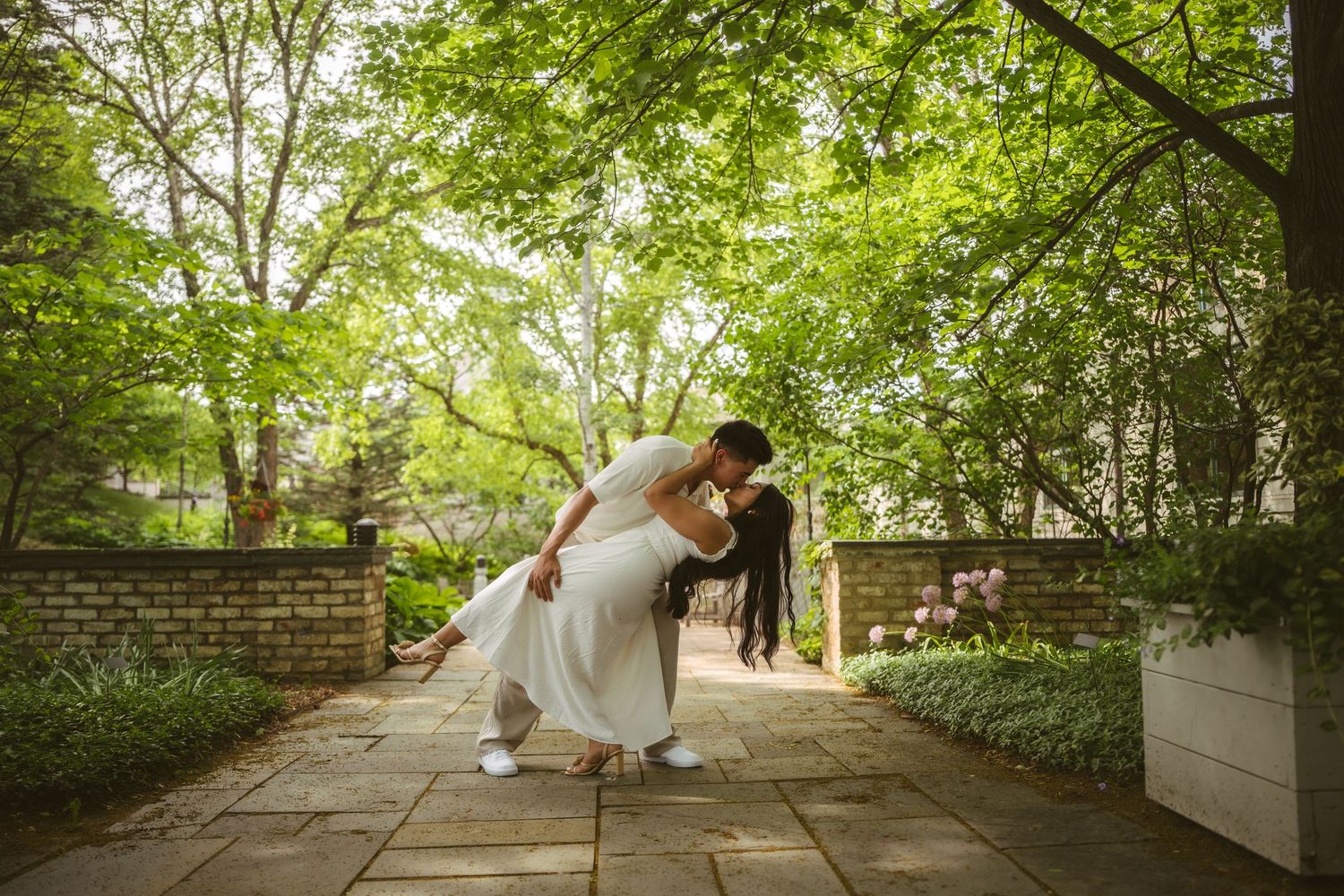 Couple sharing a romantic dip kiss on a stone pathway surrounded by lush green trees in a garden.