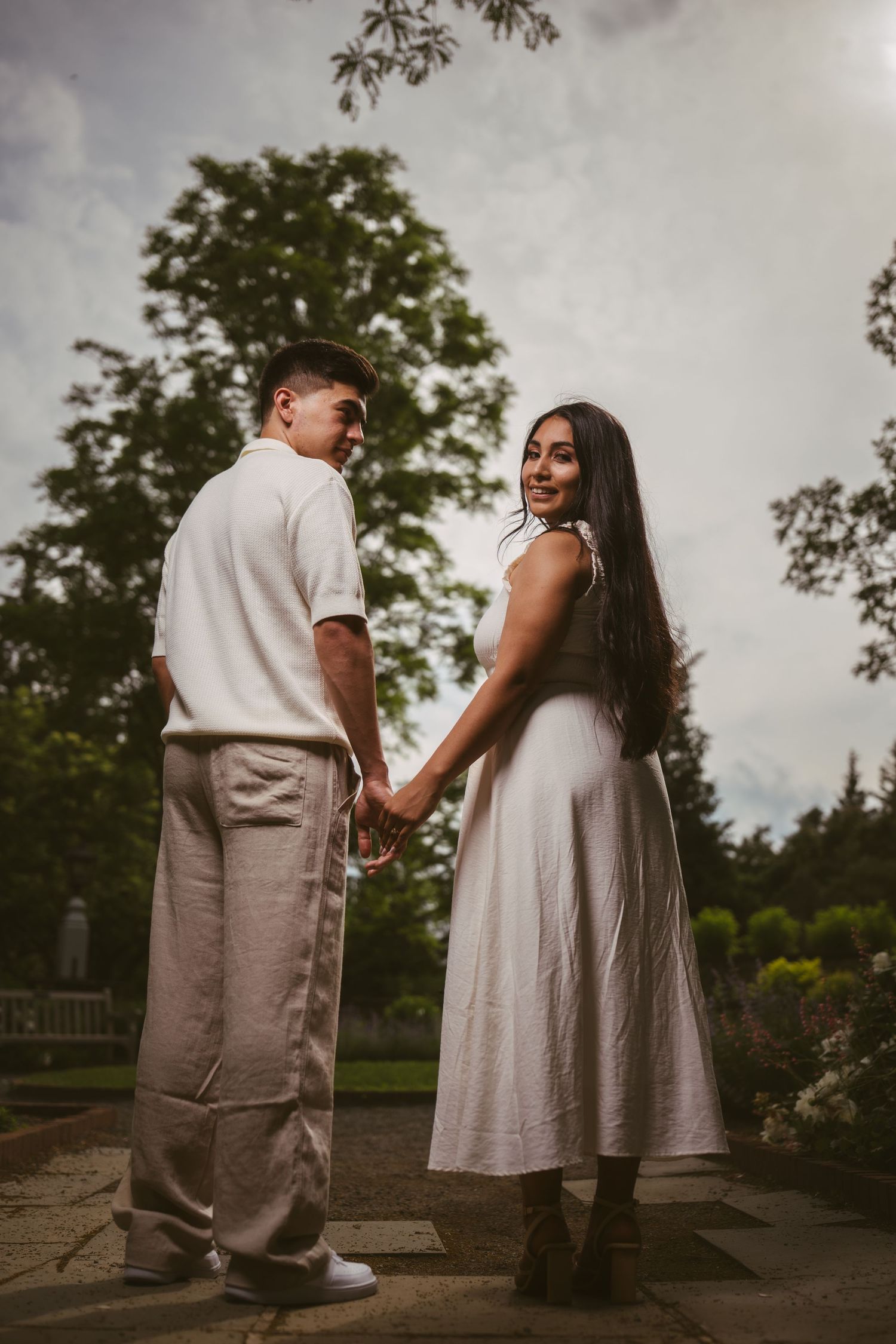 Couple holding hands in a garden, woman in white dress smiling over shoulder, man in white shirt and tan pants.