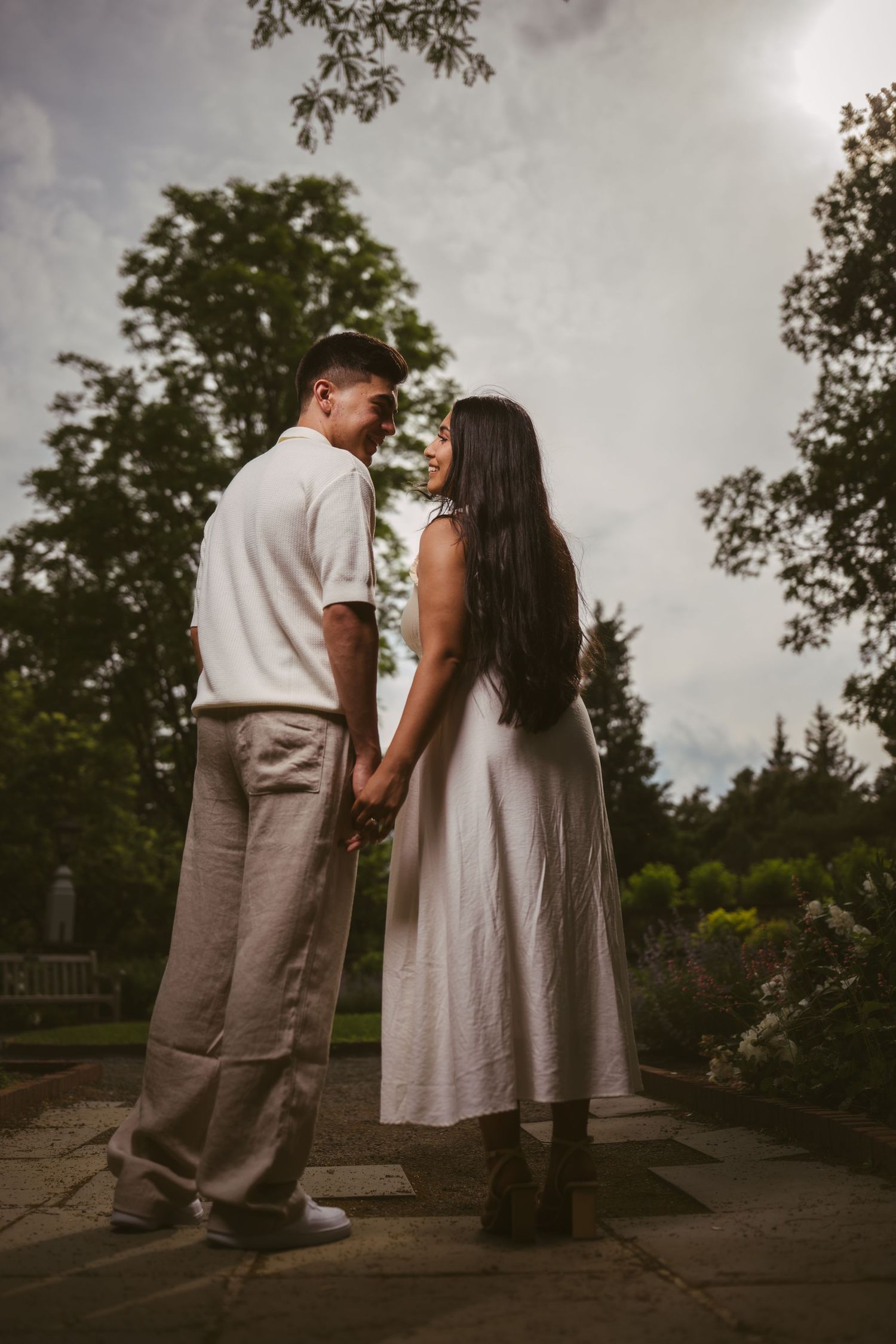 Couple holding hands in a garden, wearing white outfits, surrounded by lush green trees under a cloudy sky.