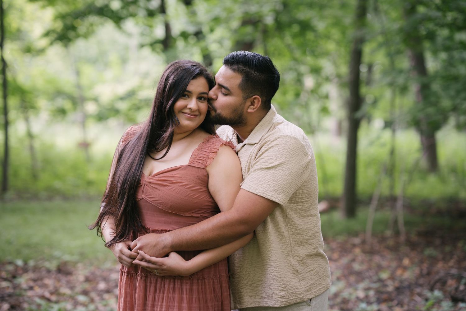 A romantic couple embraces in a wooded setting wearing coordinated pink and beige outfits.