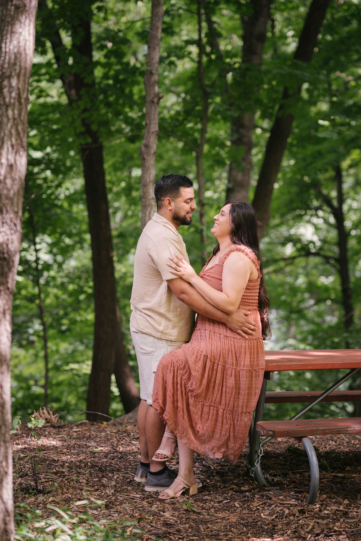 Couple shares an intimate moment by a picnic table in a lush forest setting.