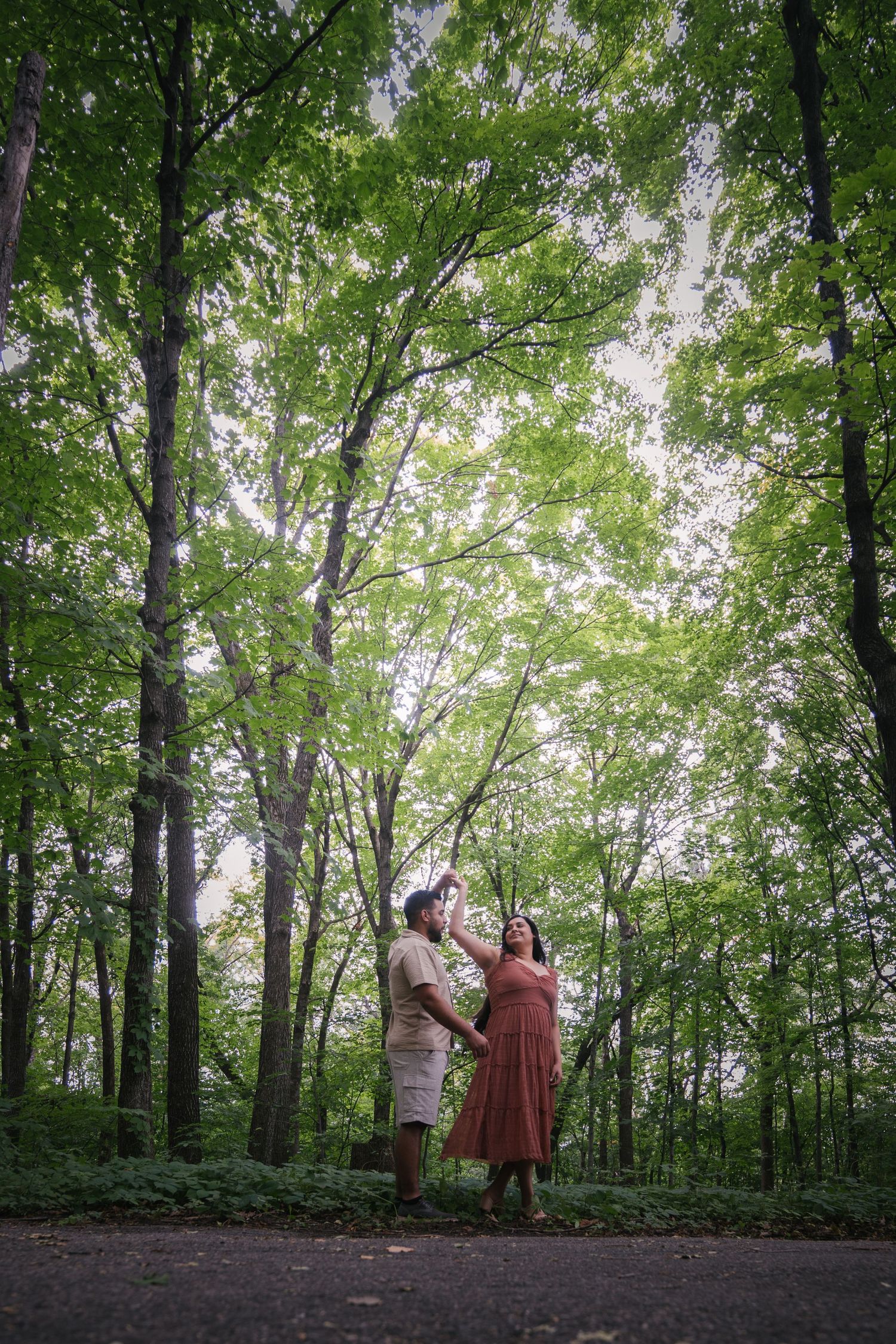 A couple stands together on a wooded trail surrounded by tall green trees.