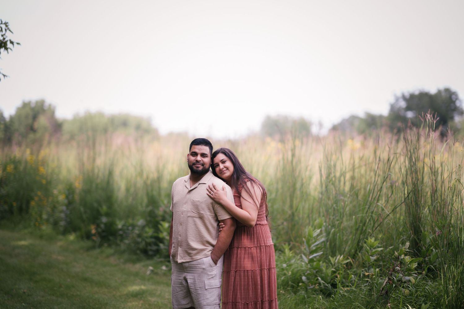 Couple poses together in a meadow with tall grass and trees in the background.