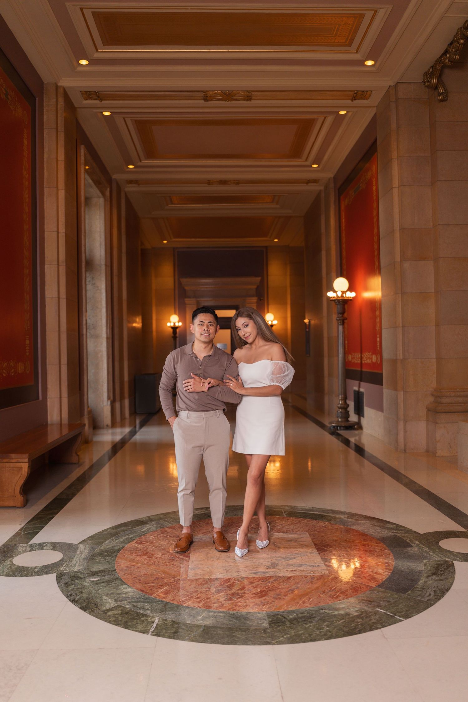 A couple poses together in an elegant hallway with ornate ceiling details and marble flooring.