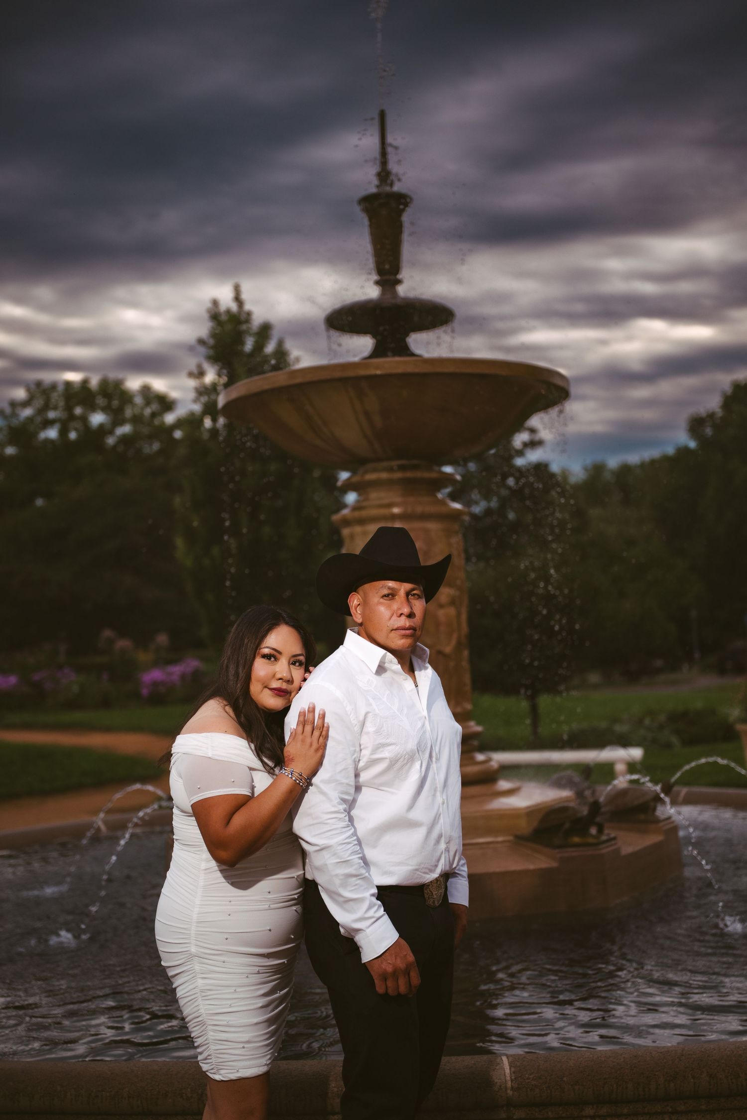A couple poses romantically by a fountain under moody storm clouds.