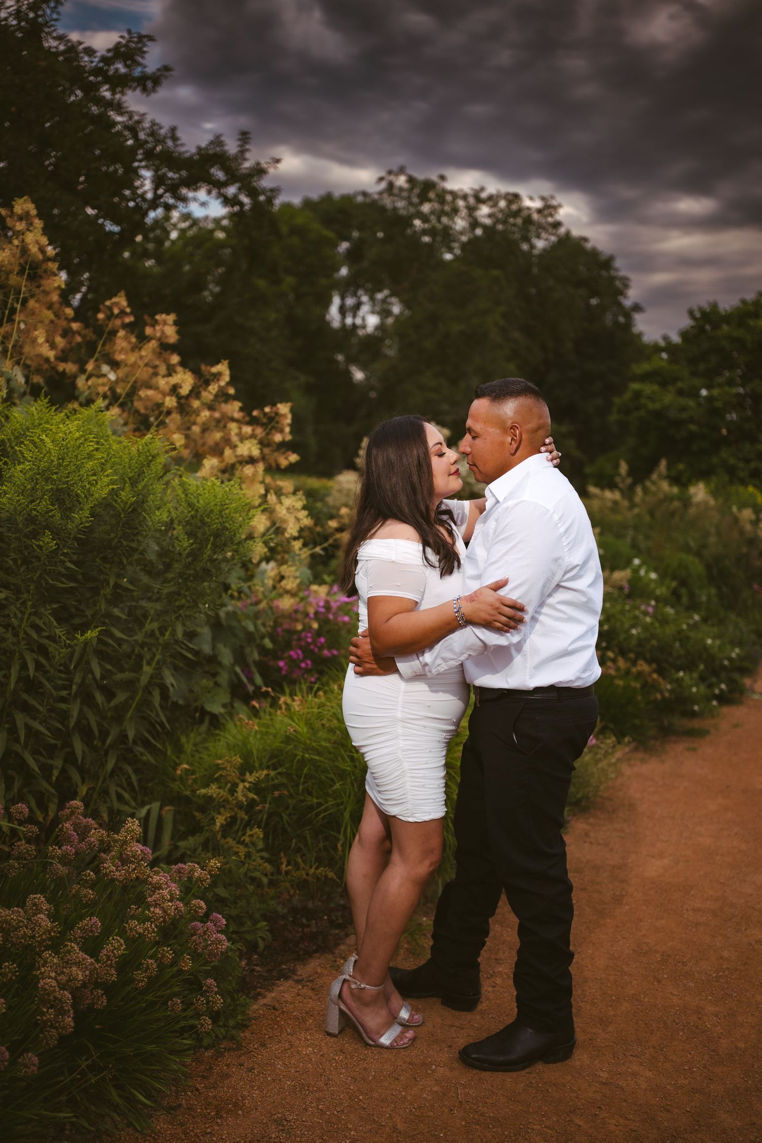 A romantic couple dances together on a dirt path surrounded by blooming flowers and trees at sunset.