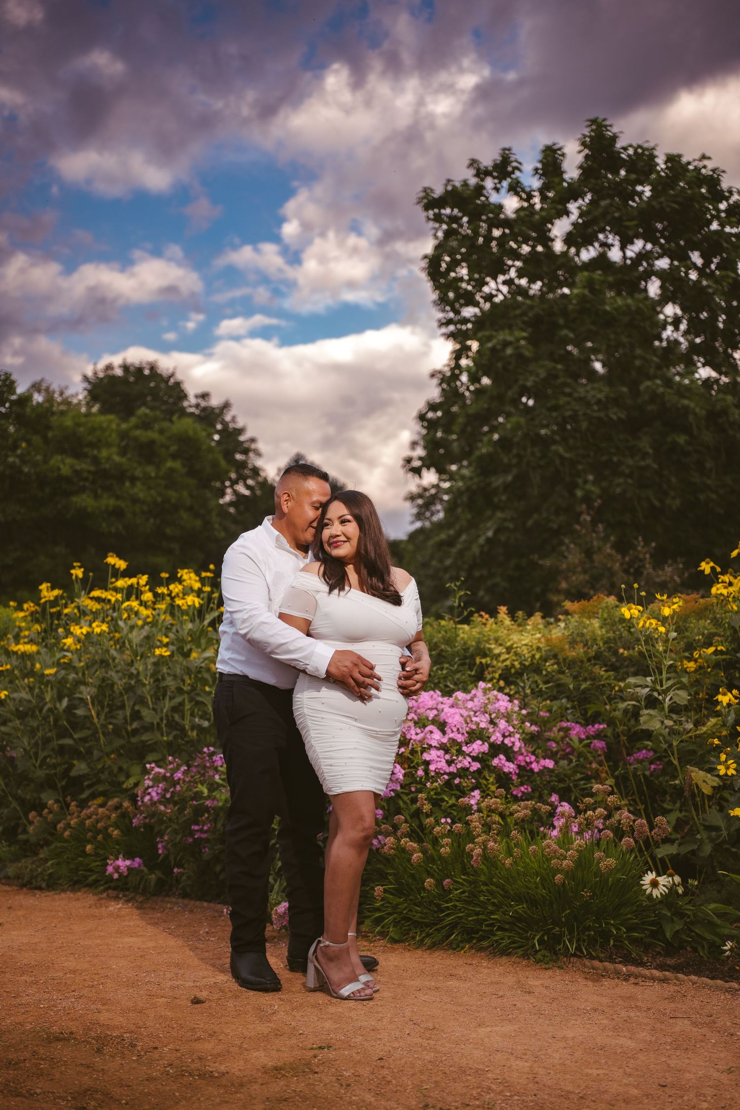 A couple shares an intimate moment in a garden filled with yellow and pink flowers under dramatic clouds.